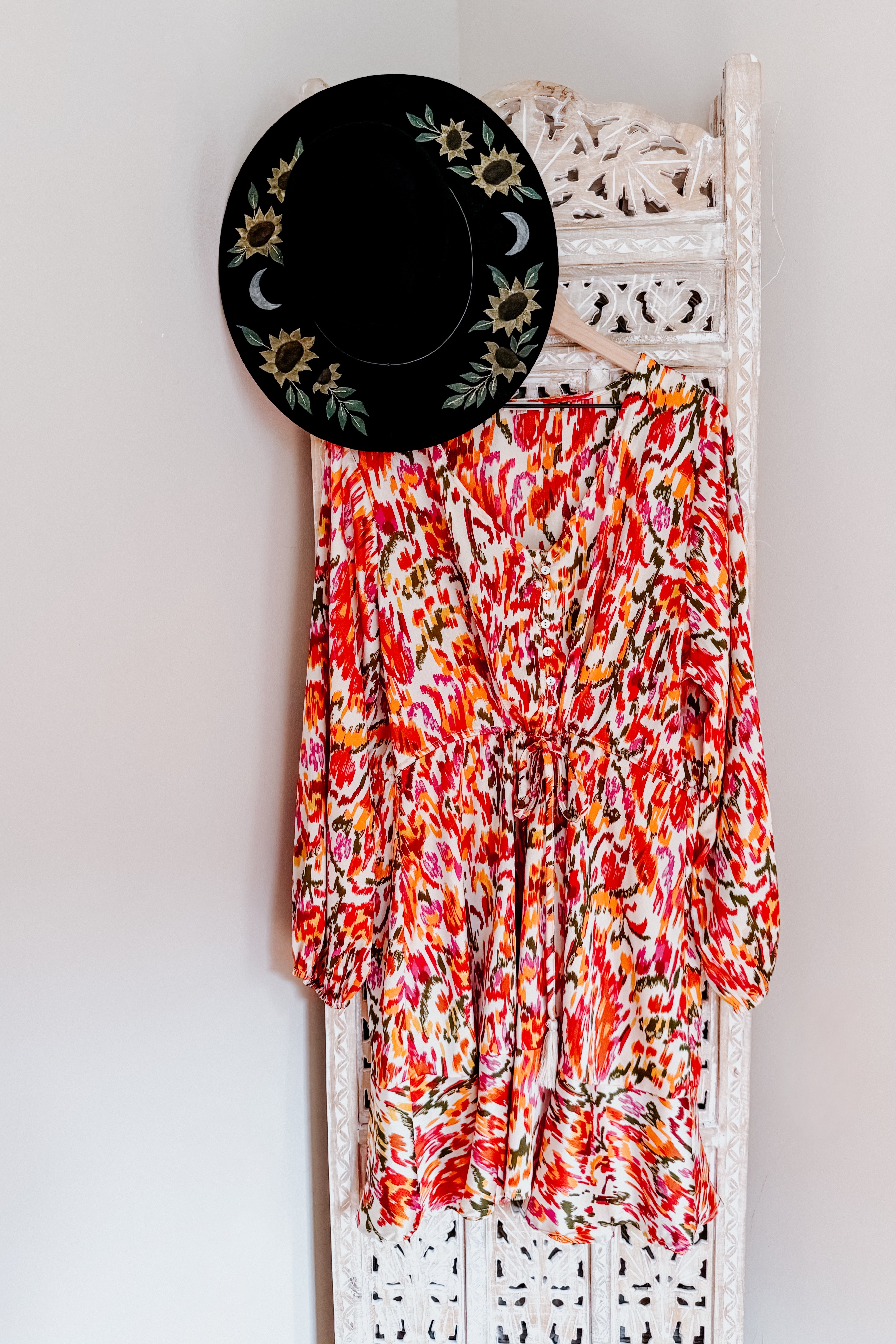 Colorful boho dress with balloon sleeves and cinched waist hanging on carved white wooden screen with black floral hat
