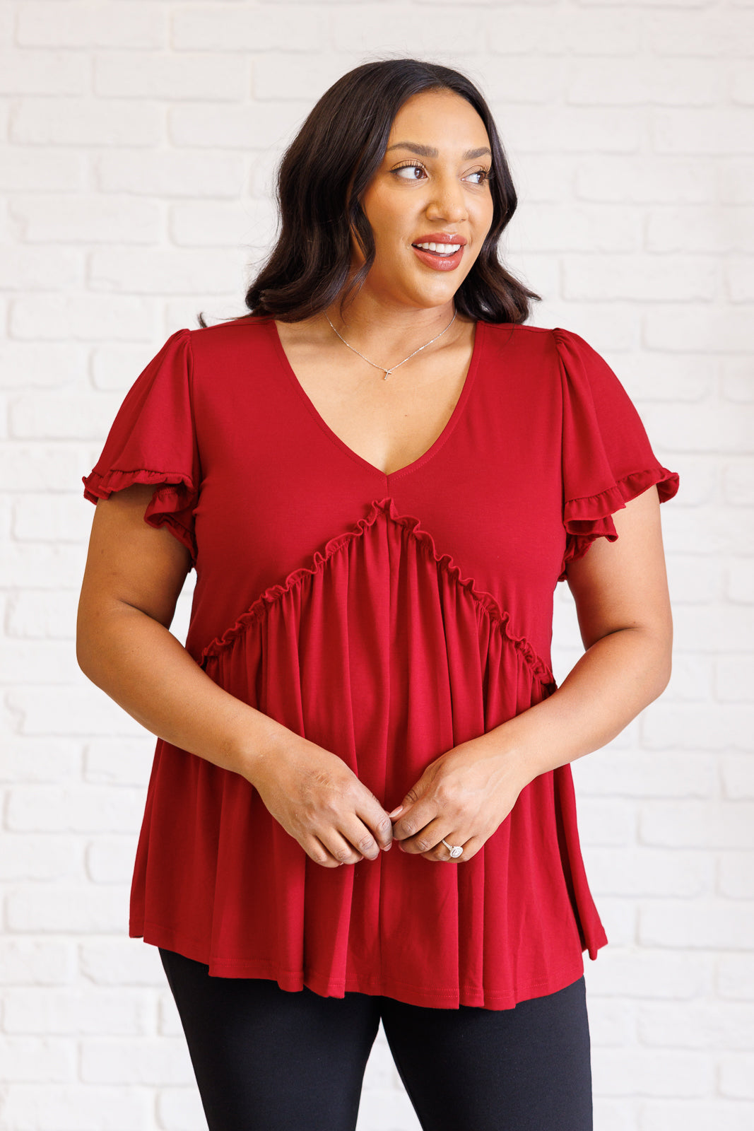 Curvy woman wearing a red V-neck top with ruffle detail and short flutter sleeves standing against white brick wall