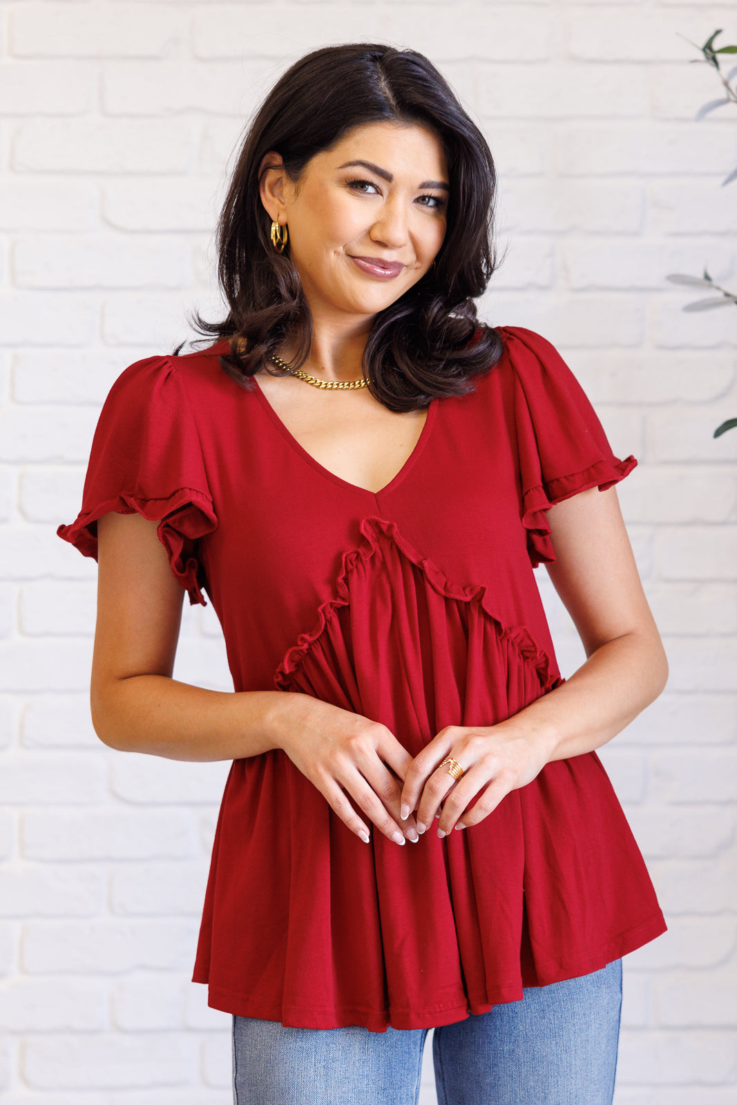 Woman wearing size-inclusive red V-neck top with ruffle details and puff sleeves against white brick wall