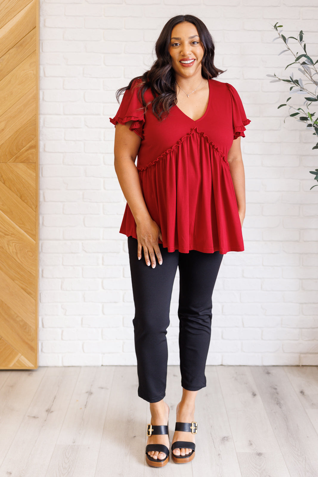 Curvy woman wearing red V-neck top with ruffle details and flutter sleeves standing in bright room with plants