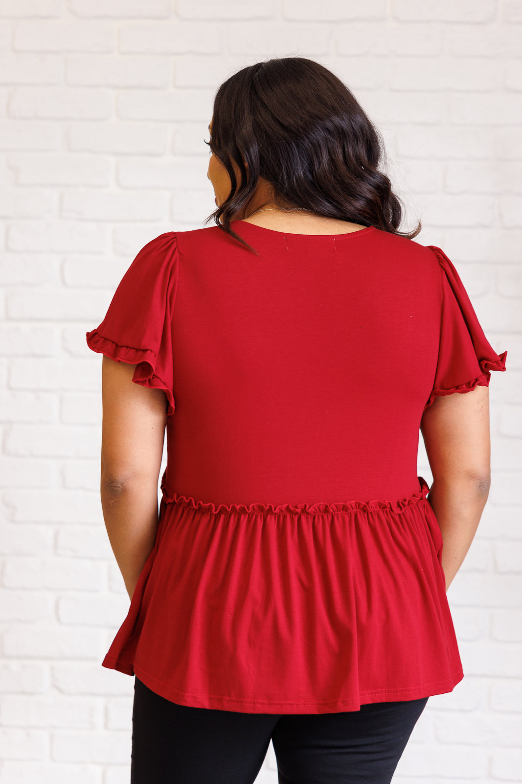 Woman wearing vibrant red ruffle detail top with flutter sleeves styled with black pants against white brick wall