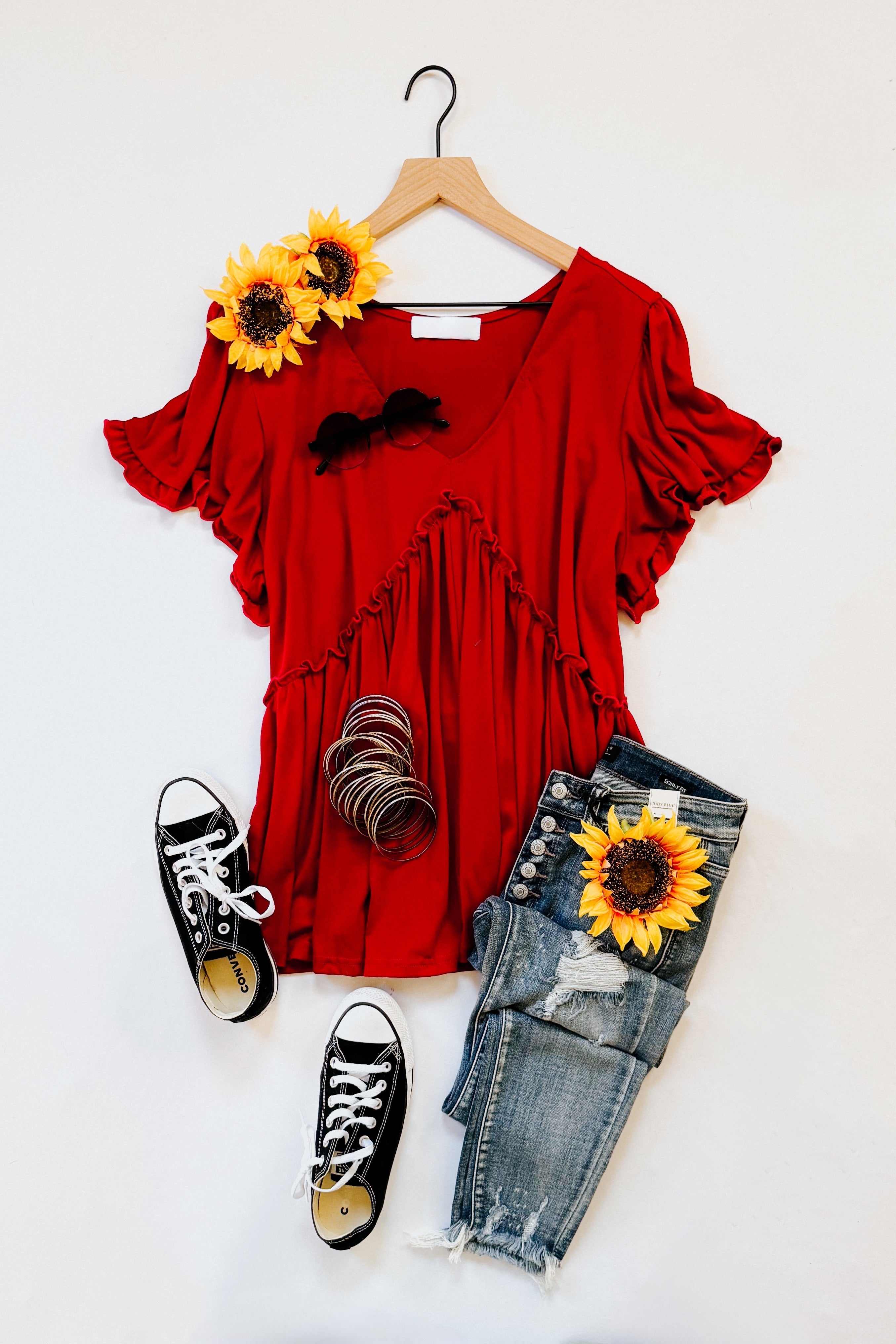 Red V-neck top with ruffle detail paired with distressed blue jeans, black sneakers, and sunflower accents on hanger