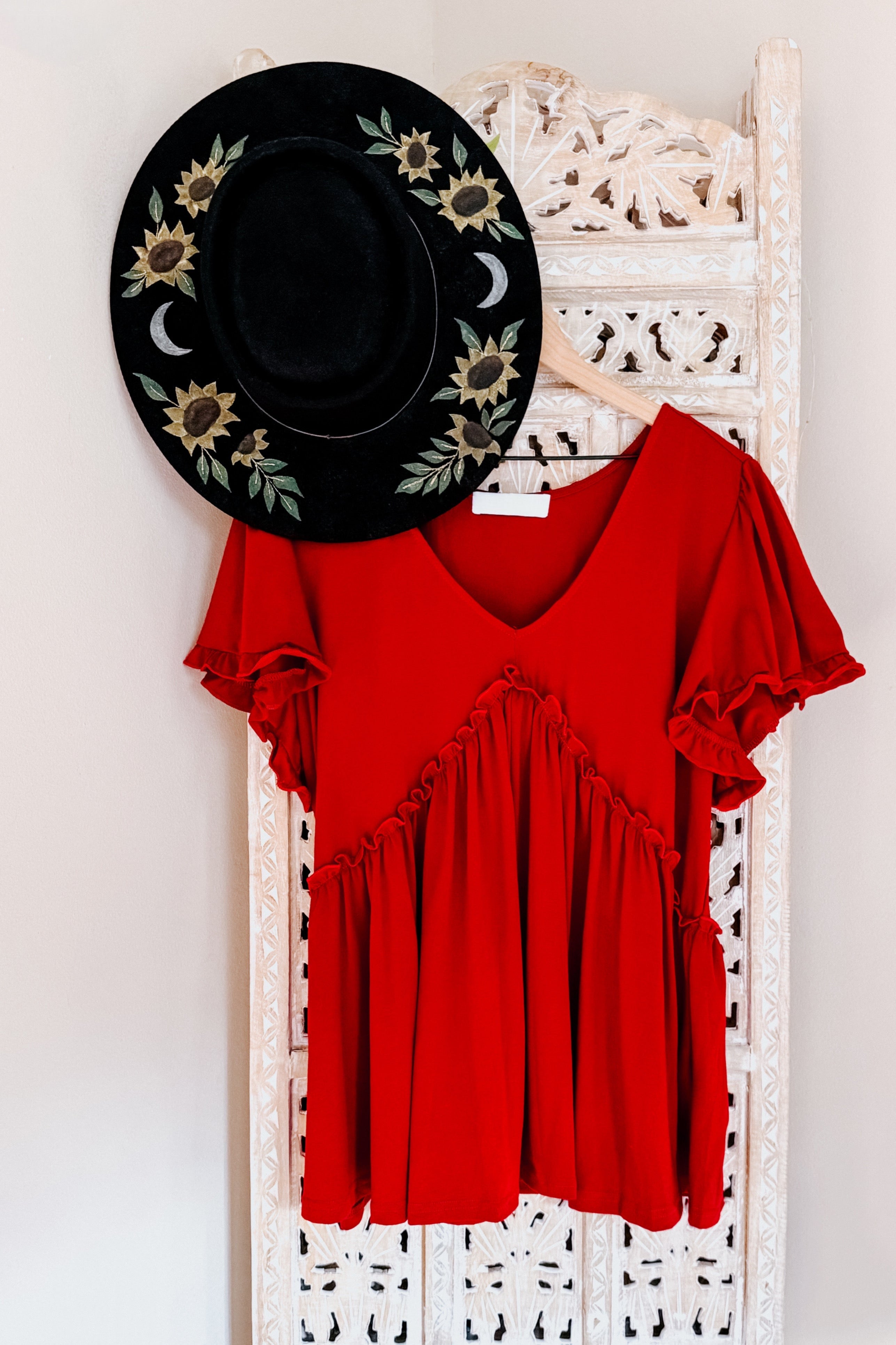 Red V-neck top with ruffle detailing and puffed sleeves hanging next to black hat with sunflower embroidery