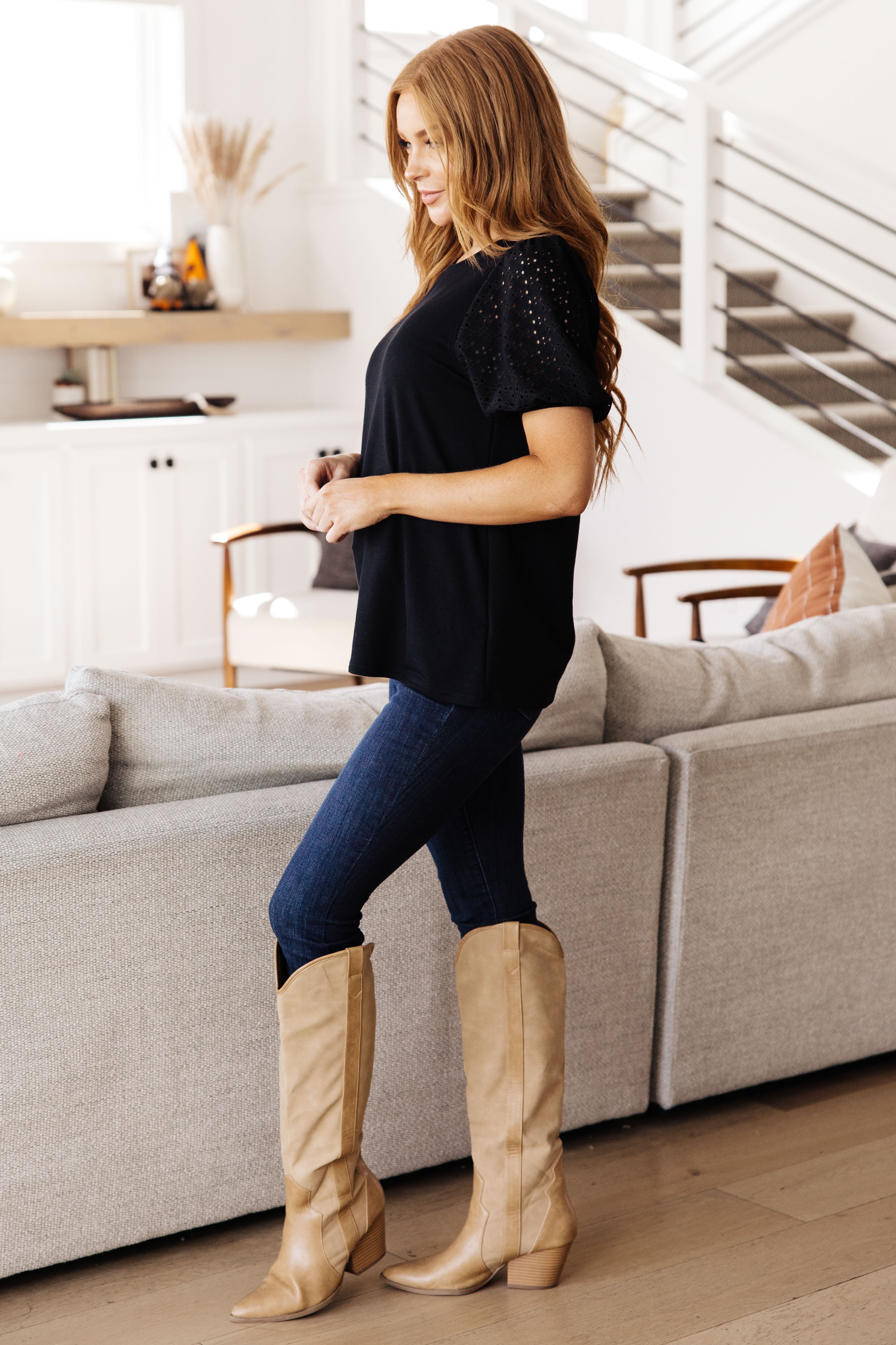 Woman wearing a black top with lace sleeves paired with dark skinny jeans and tan knee-high boots in cozy living room