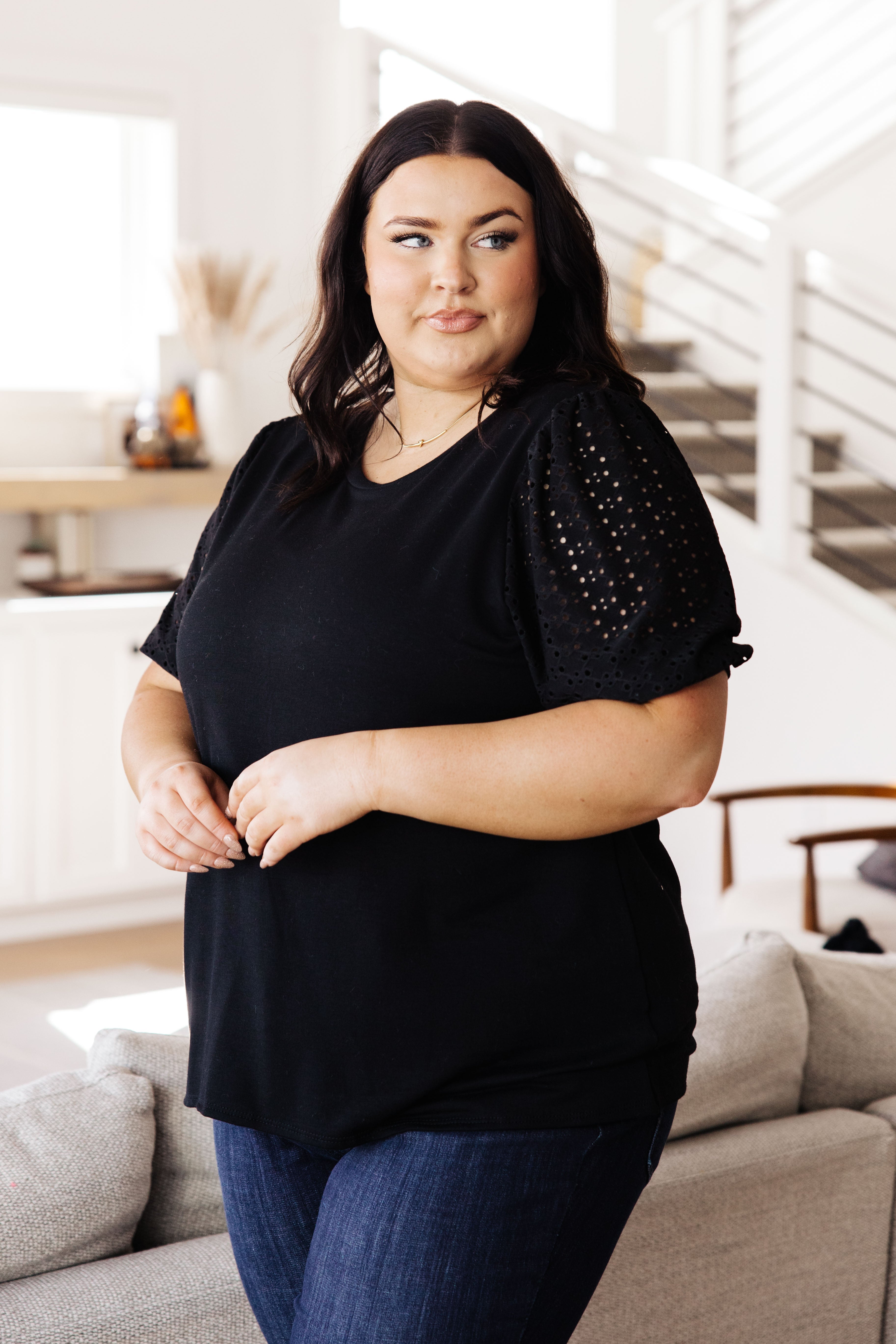 Plus size woman wearing a black top with eyelet lace puff sleeves paired with dark blue jeans in a bright casual room
