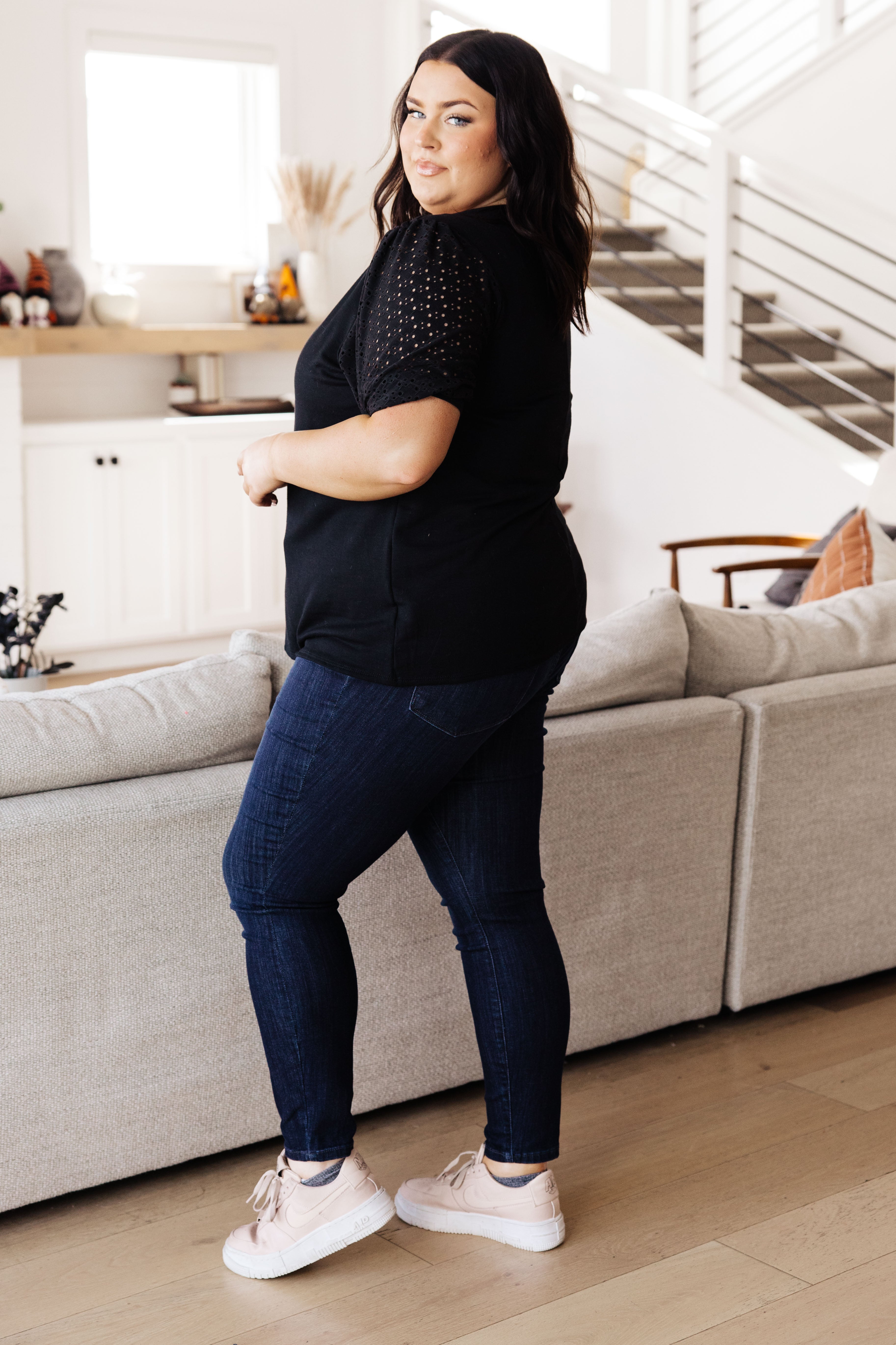 Plus size woman wearing black lace sleeve top with dark blue jeans and light sneakers in cozy modern living room