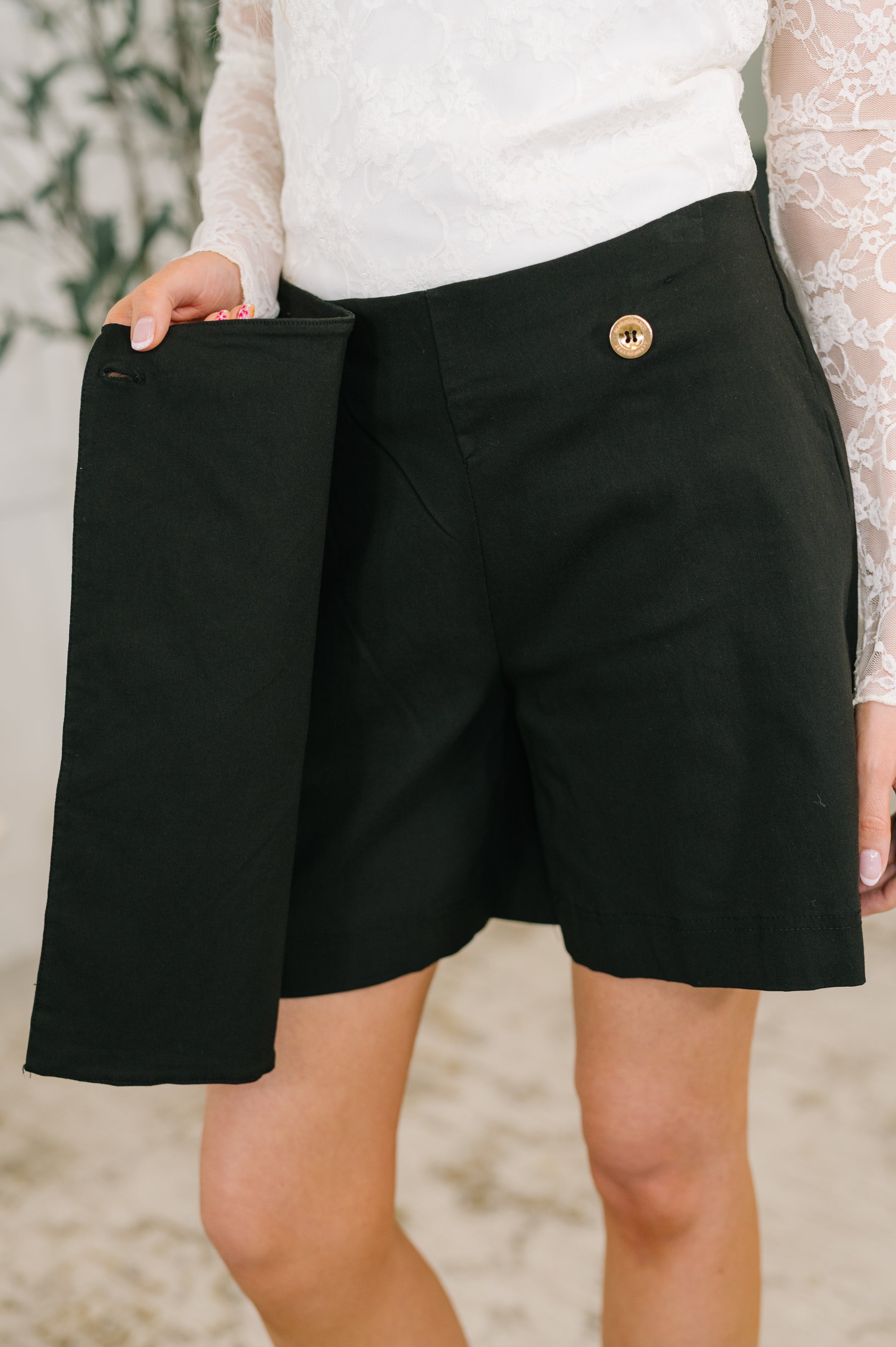 Close-up of black faux wrap skort with wooden button detail held open by woman wearing white lace top indoors