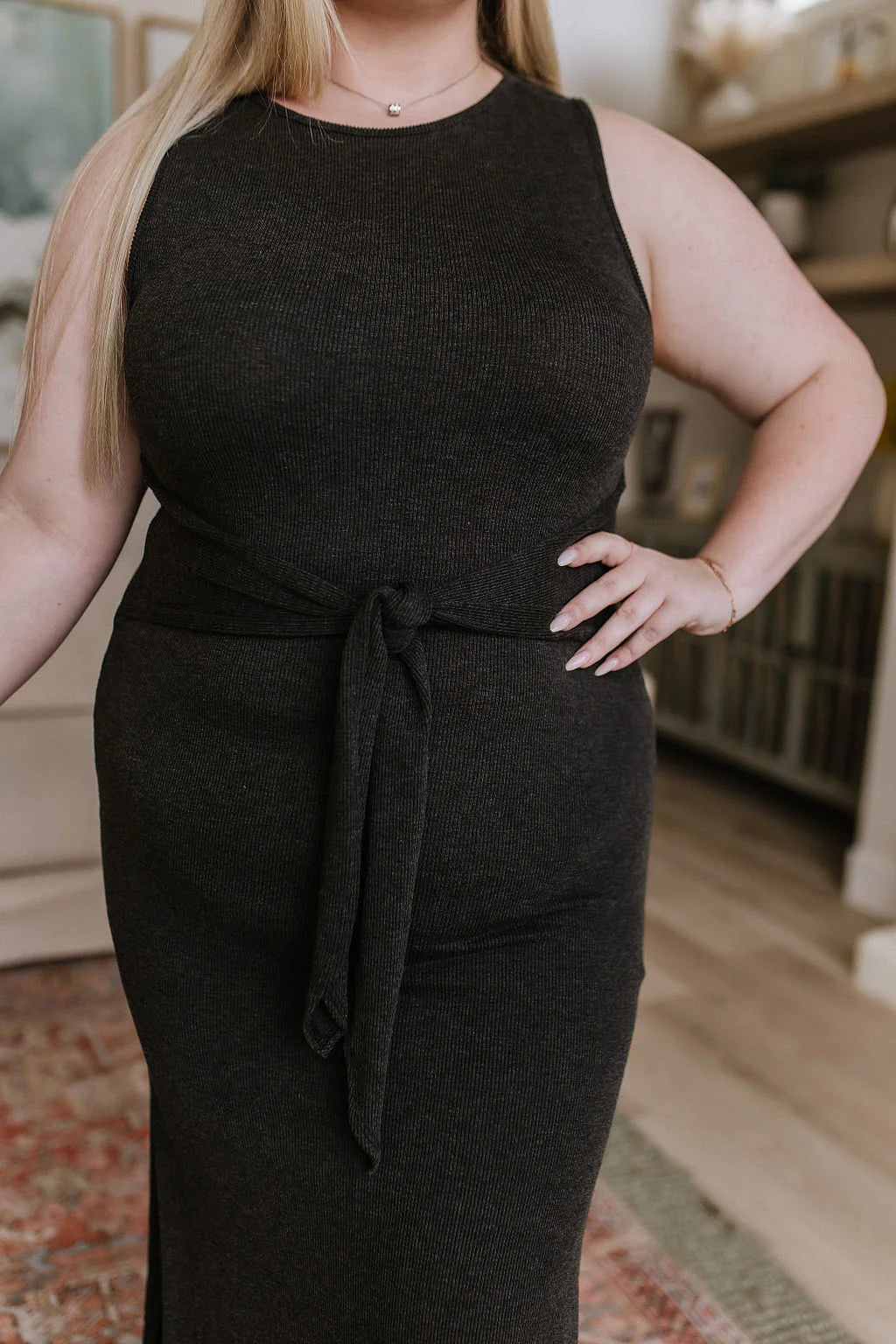 Curvy woman wearing charcoal knit midi dress with waist tie detail standing indoors in a stylish home setting