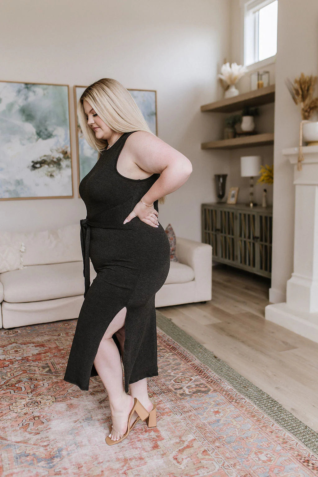 Curvy woman in a charcoal sleeveless midi dress with side slit and waist tie, wearing nude block heels indoors