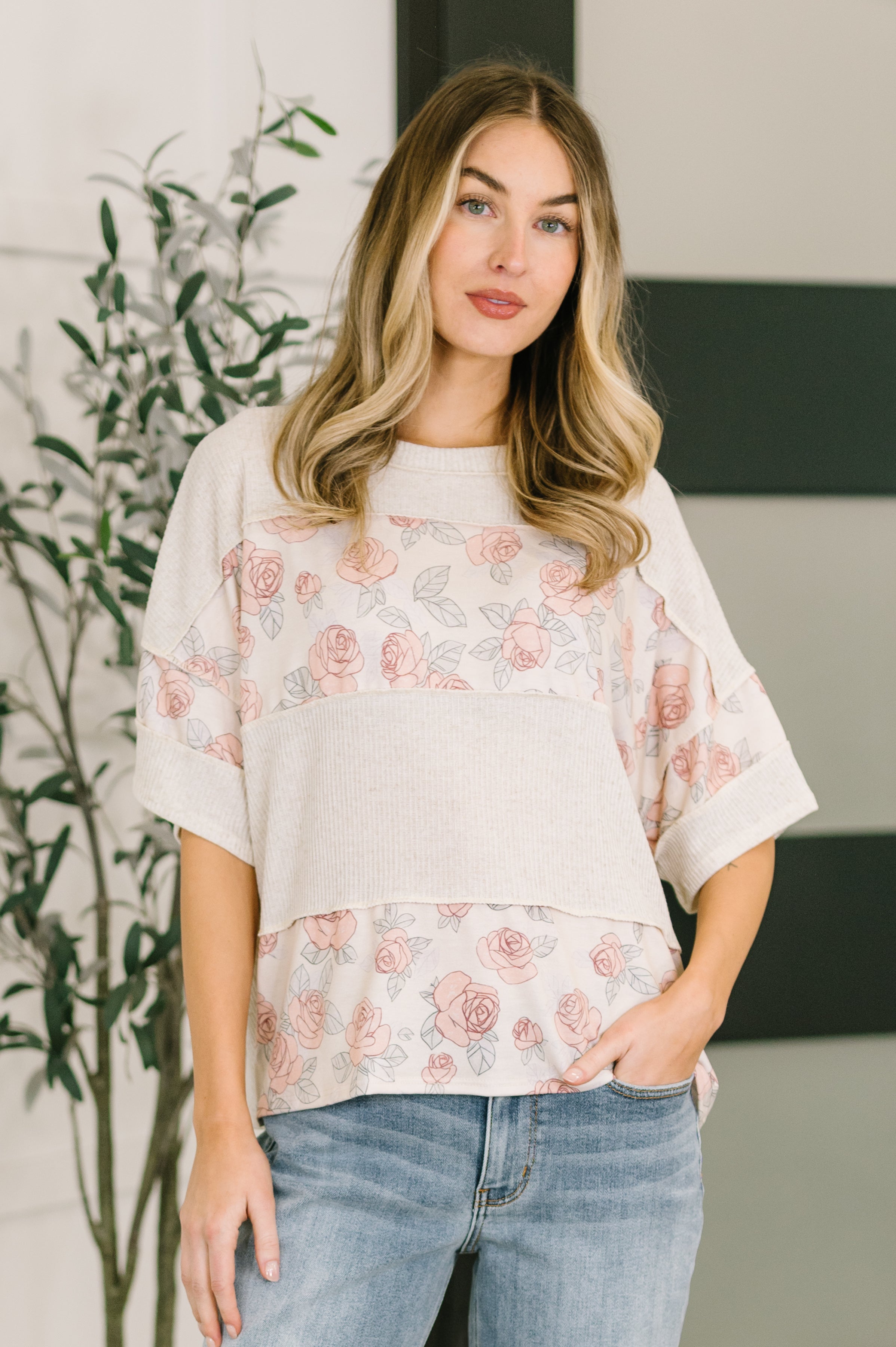 Woman wearing relaxed pink floral color block top with ribbed knit and rolled sleeves paired with blue jeans indoors