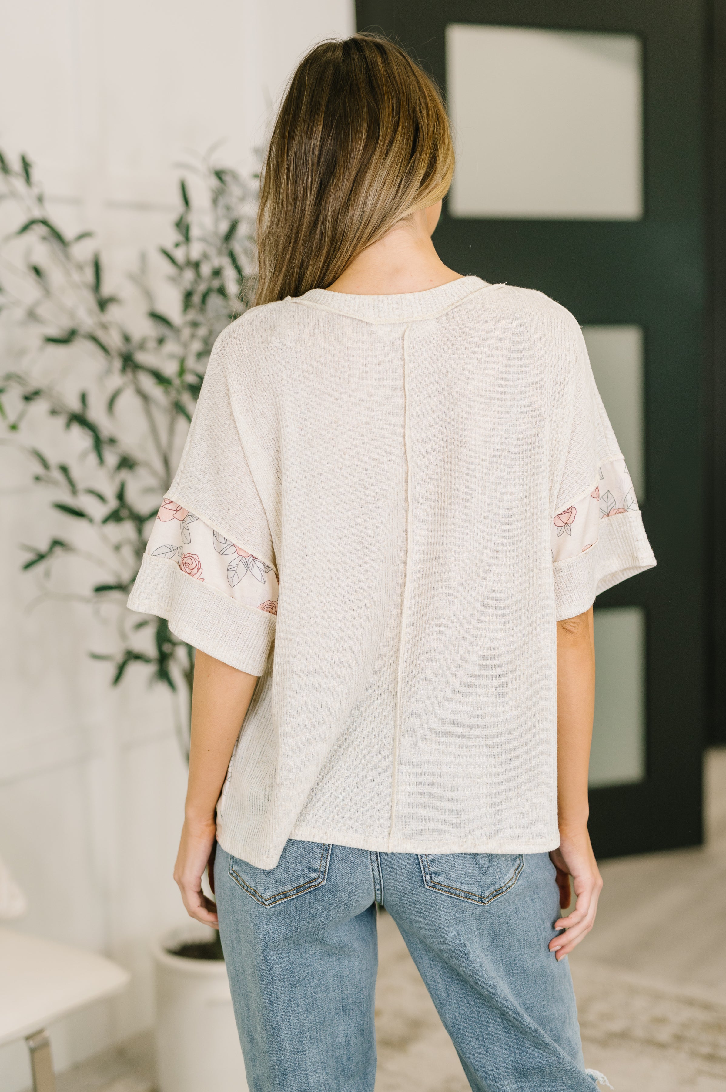 Back view of relaxed off-white top with wide sleeves featuring subtle pink rose pattern and casual light blue jeans indoors