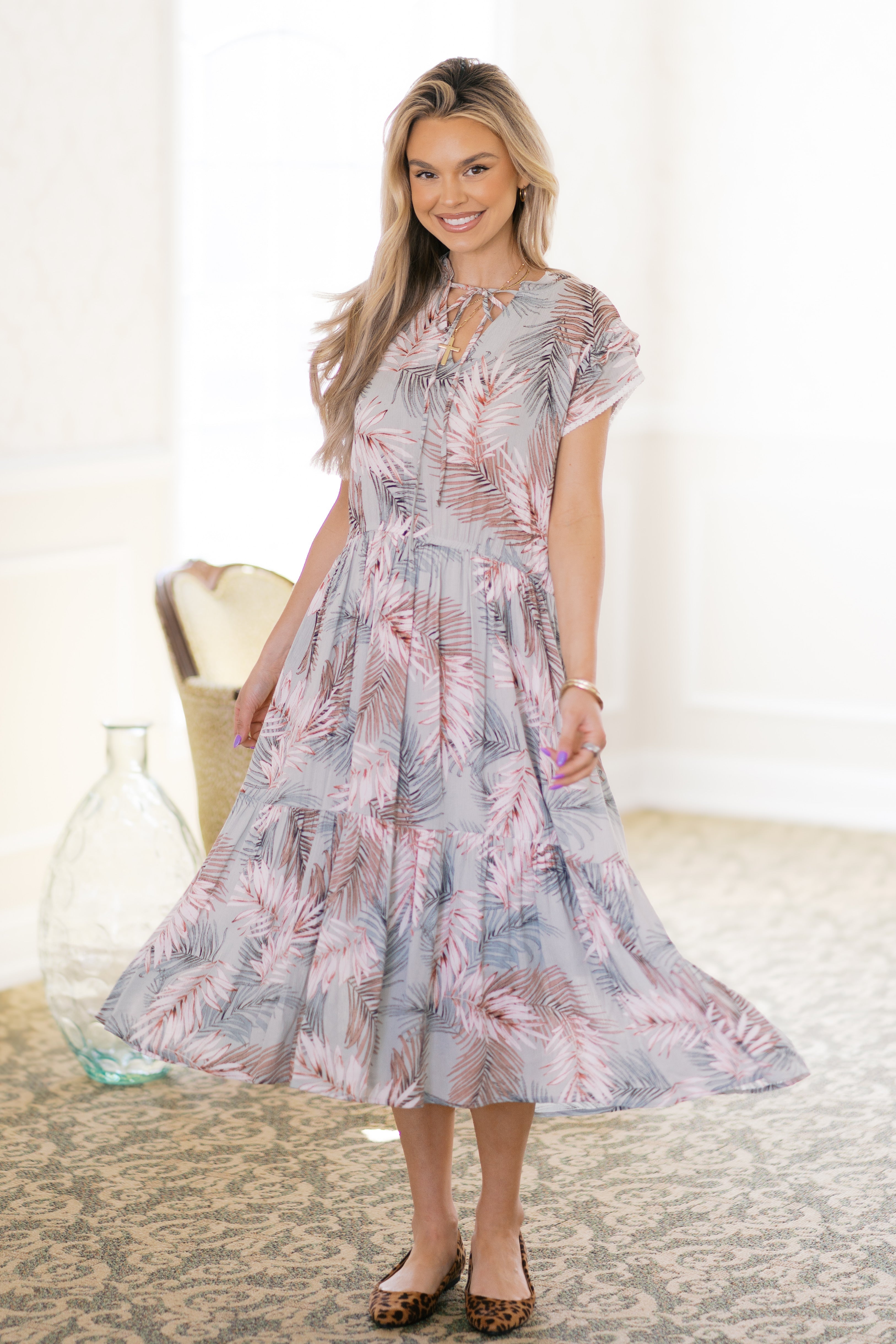 Smiling woman in green tiered midi dress with palm leaf pattern and leopard print flats standing indoors