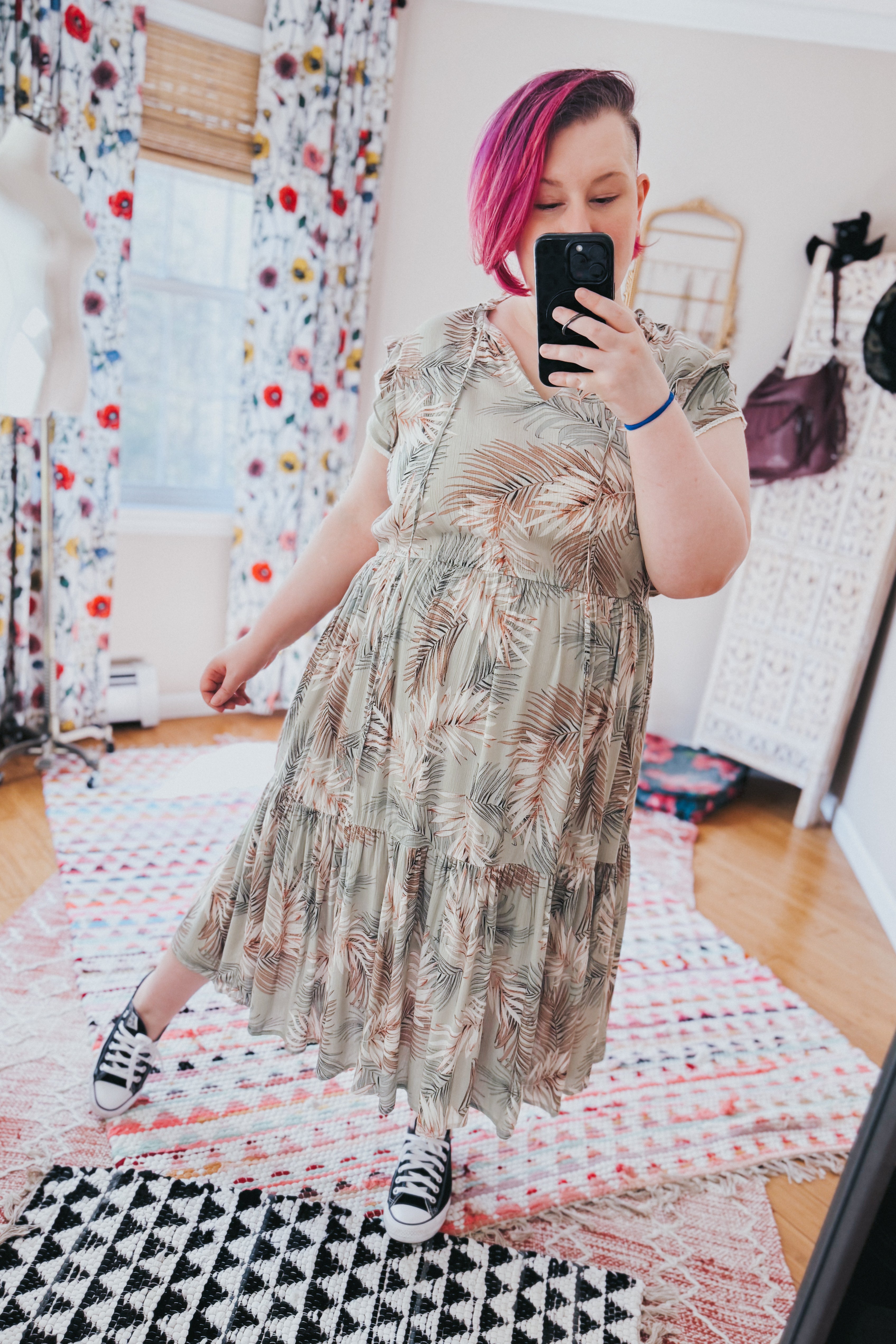 Woman with vibrant pink hair wearing a green tiered midi dress with beige palm leaf design and black sneakers indoors
