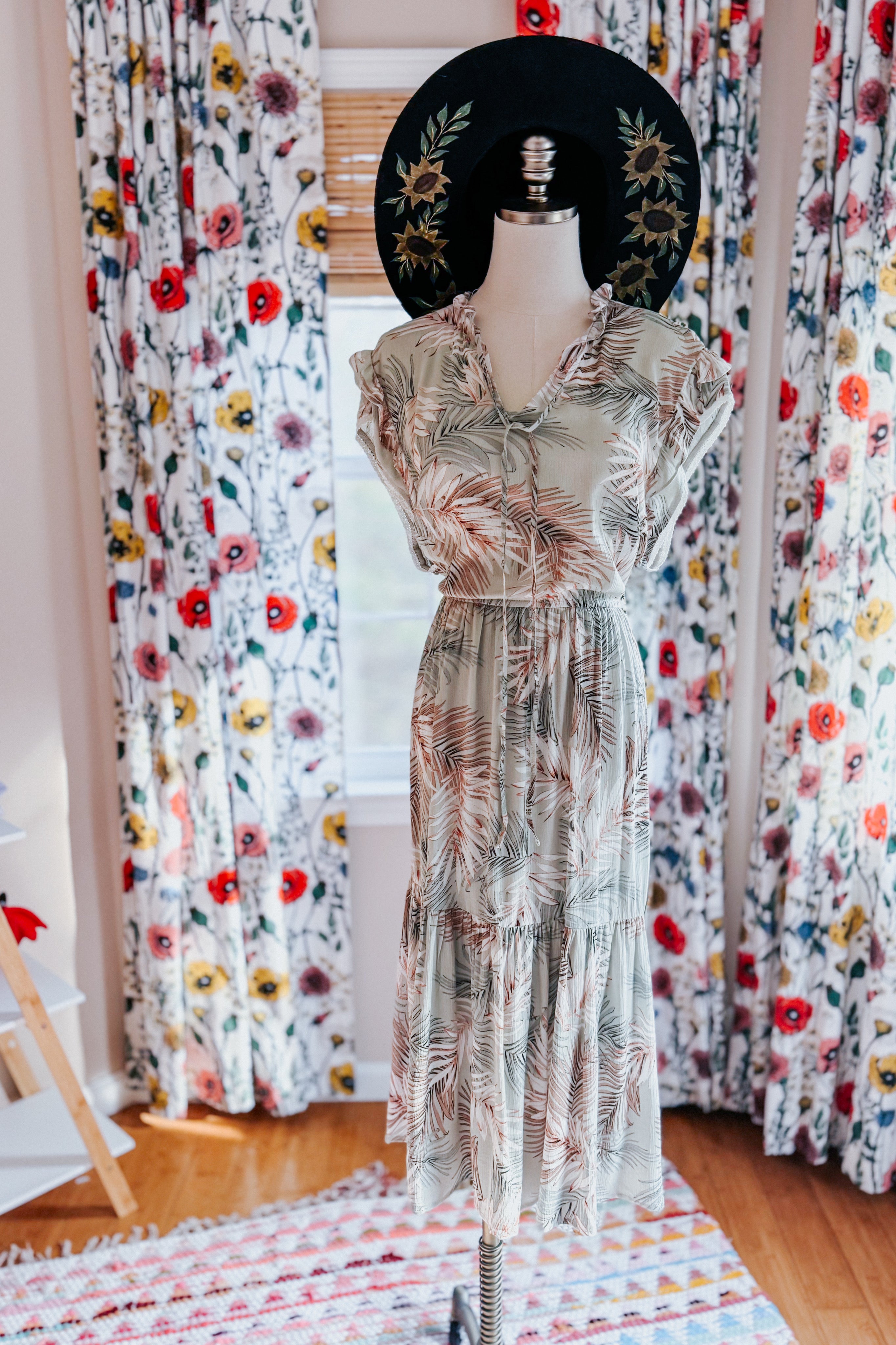 Sleeveless tiered midi dress with soft palm leaf print paired with wide-brimmed black hat in floral room setting