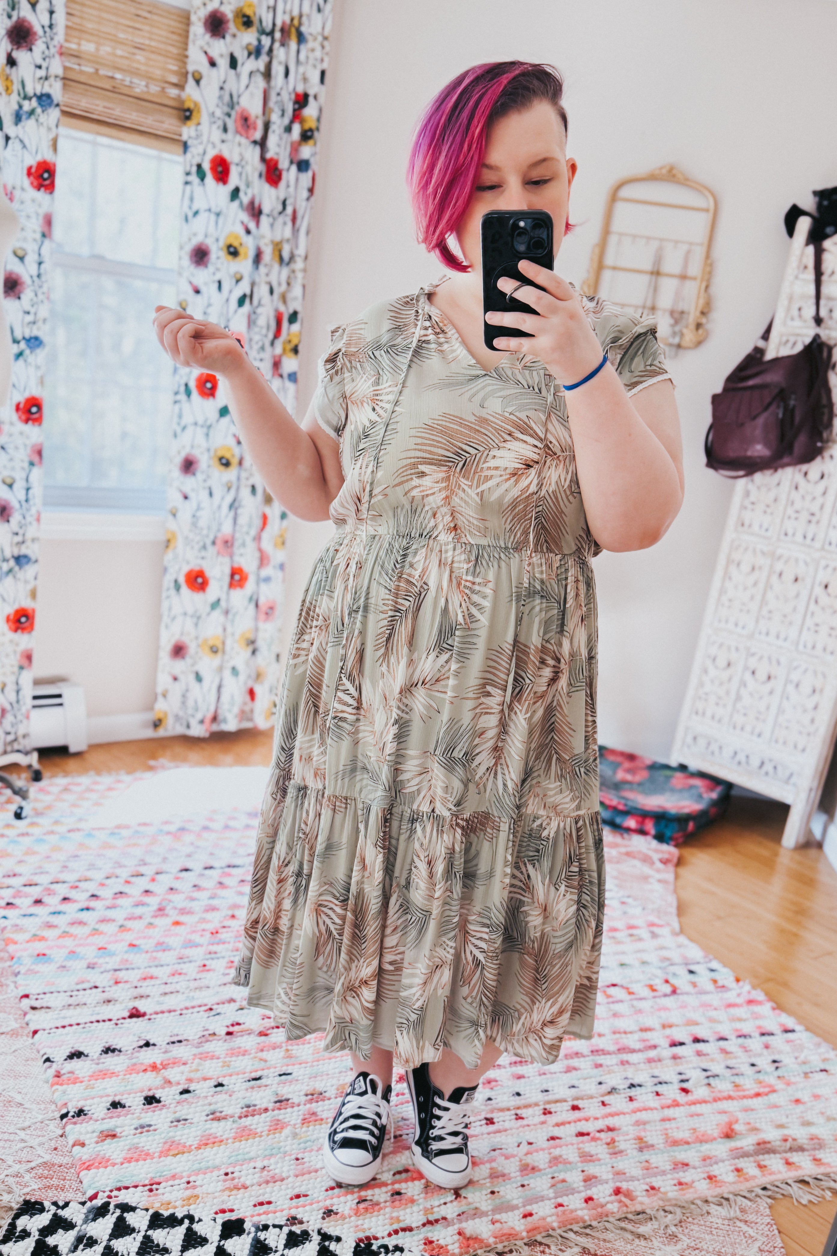 Woman with short pink hair wearing a tiered midi dress with palm leaf print and black sneakers in bright room with floral curtains