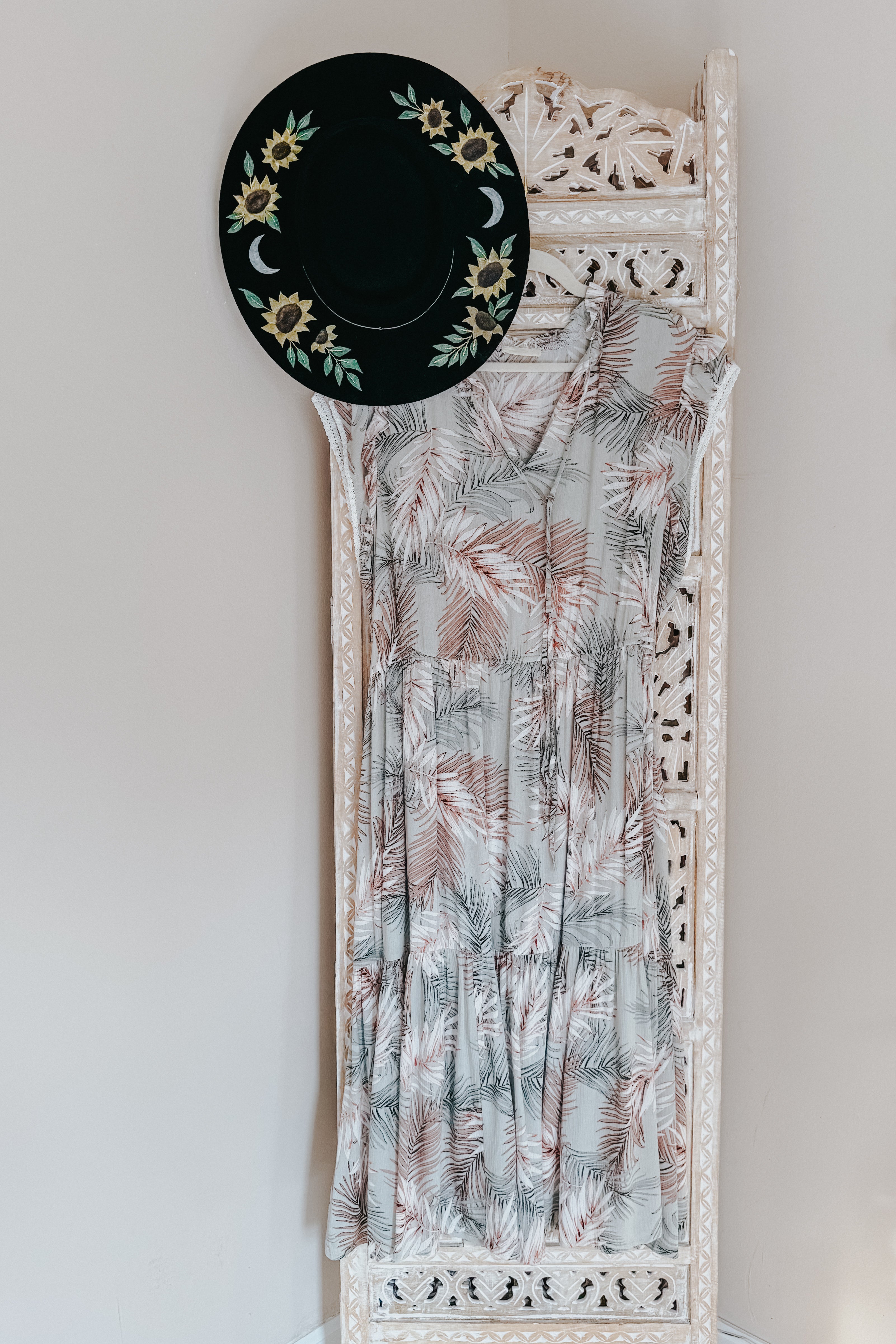 Green tiered midi dress with palm leaf pattern hanging on carved wooden screen paired with black floral hat