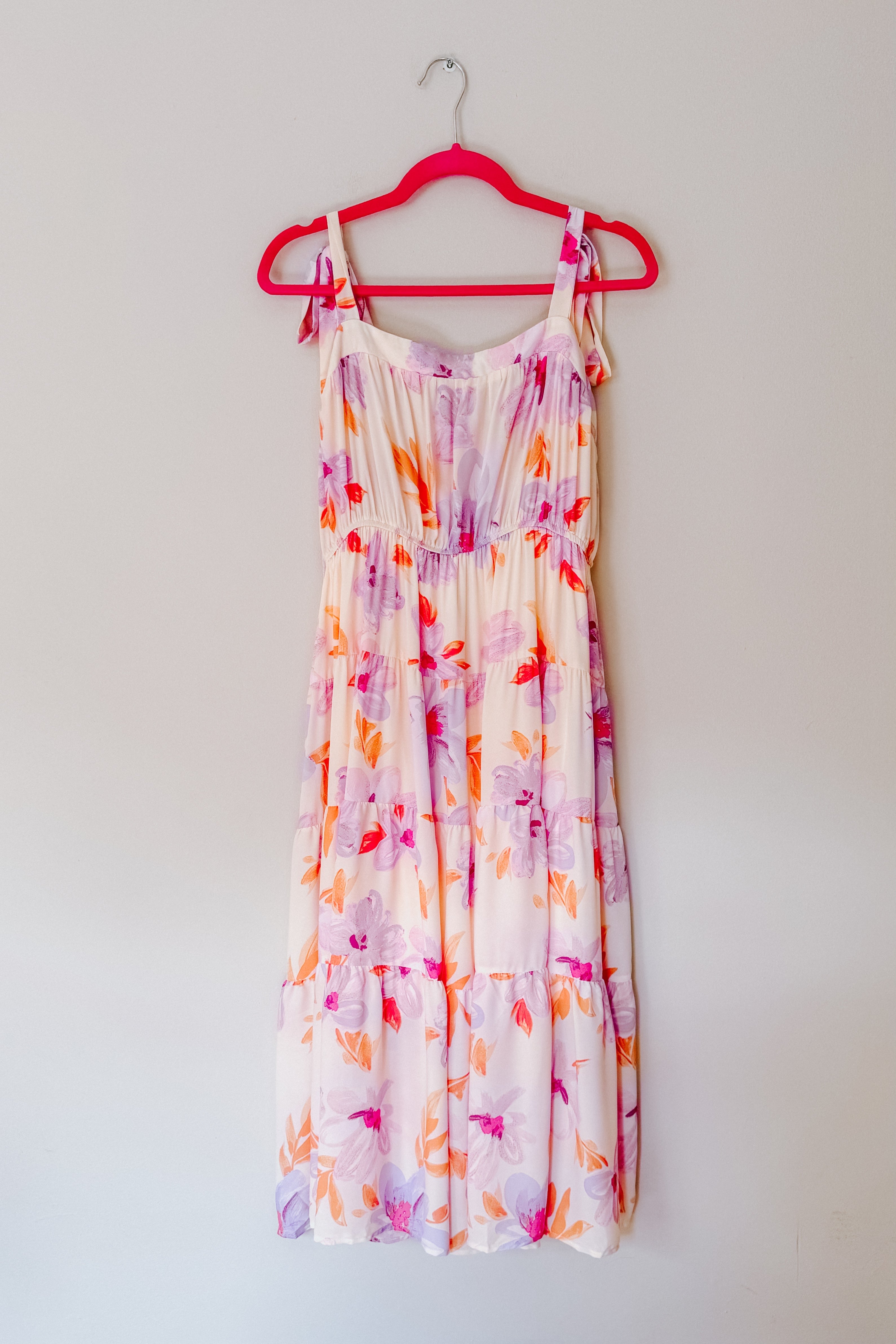 Light pastel floral midi dress with tie shoulder straps hanging on bright pink hanger against plain wall