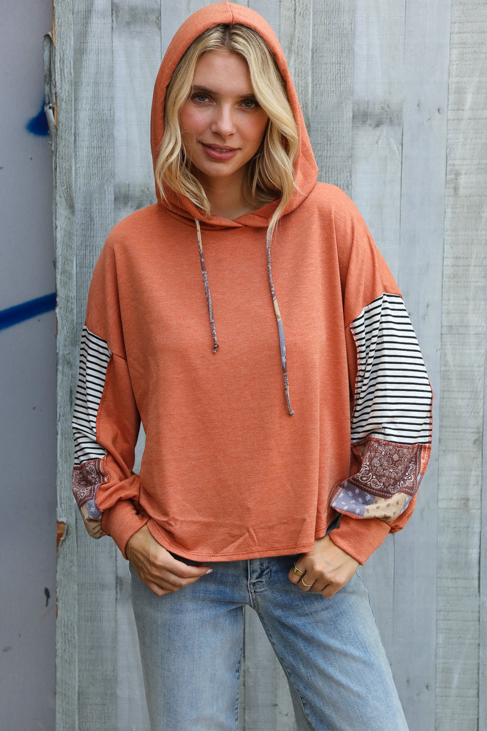 Woman in rust hoodie with striped and patchwork sleeves standing against weathered wood wall smiling confidently