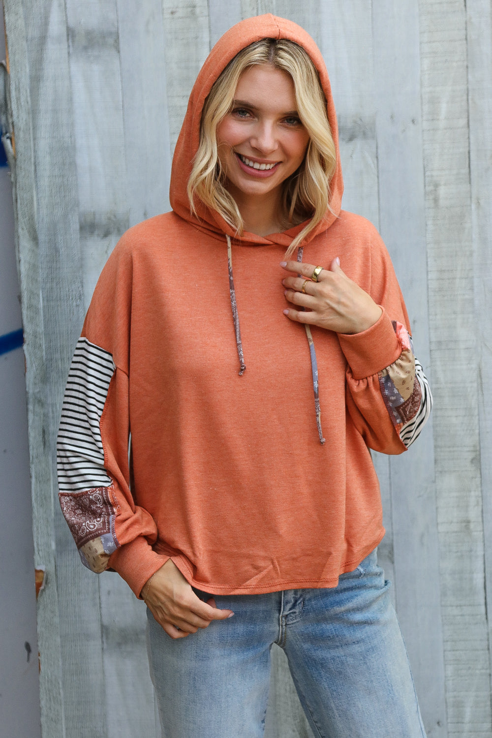 Smiling woman wearing rust hoodie with striped and patchwork sleeves and hood posing against weathered wood background