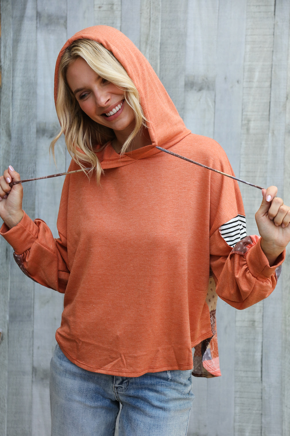 Blonde woman smiling wearing rust-colored hoodie with playful patch and striped sleeve details pulling on hood strings