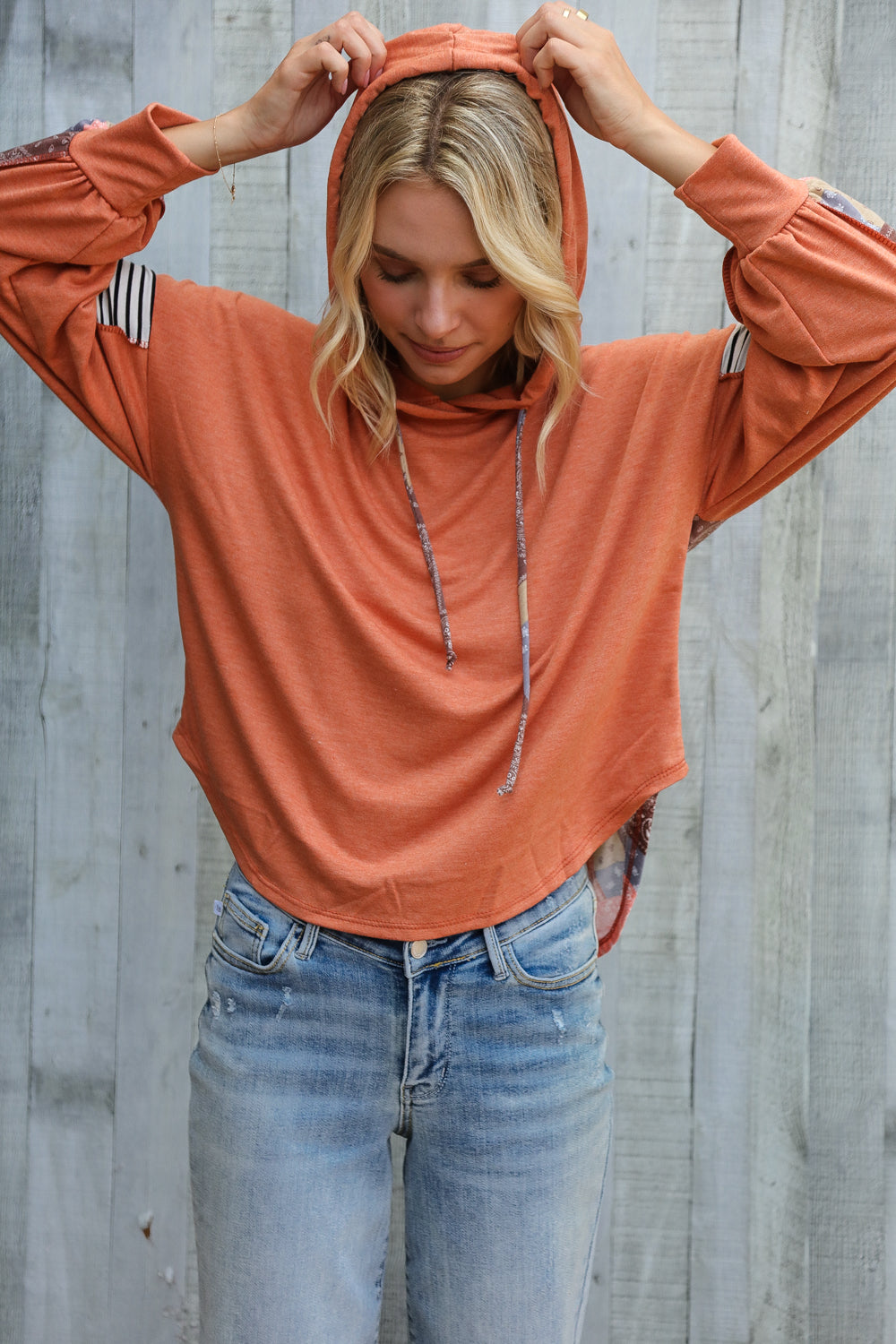 Woman adjusting hood of rust patchwork French terry hoodie with striped sleeve details and relaxed fit jeans