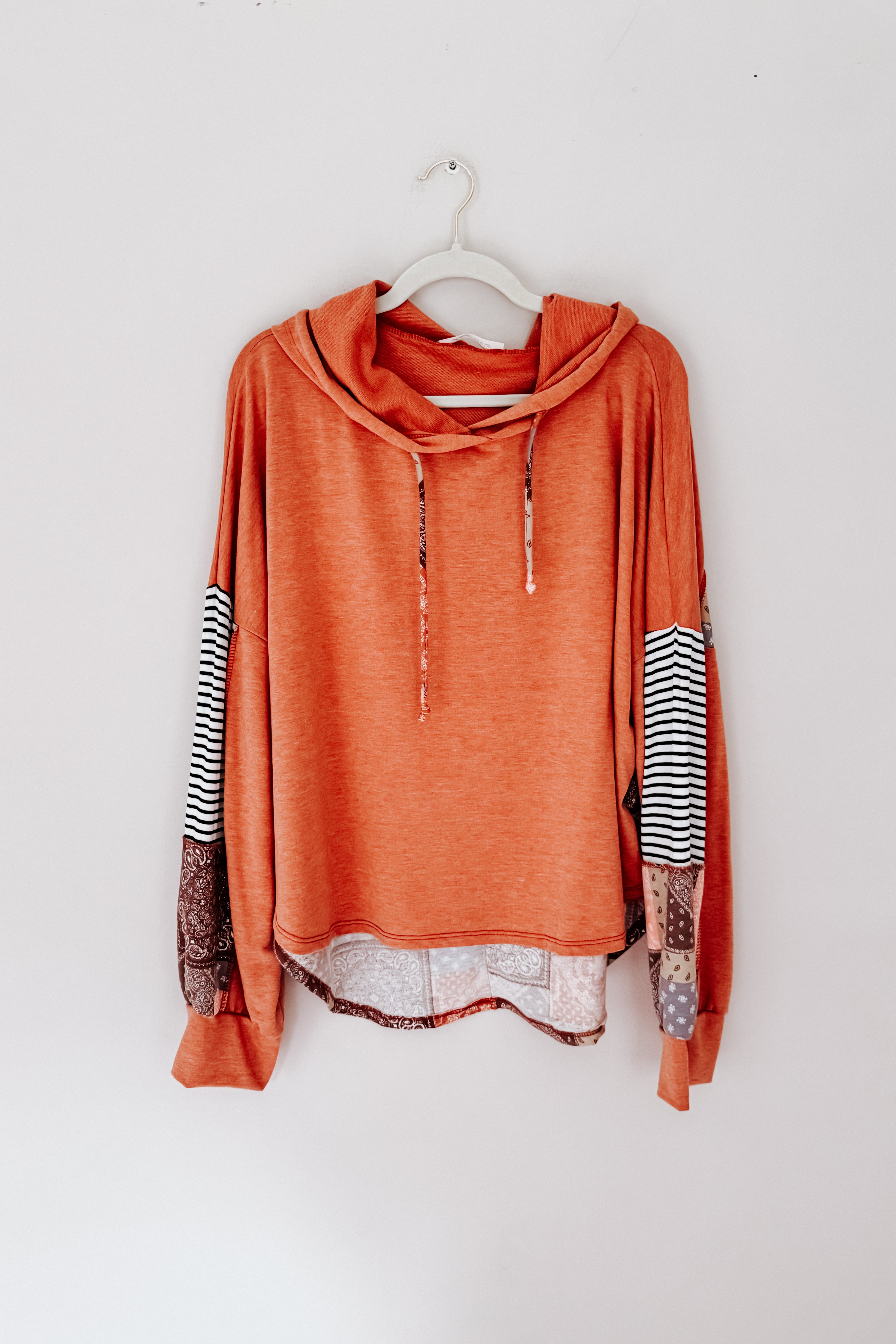 Rust-colored eclectic hoodie with patchwork elbow details and striped sleeve accents hanging on white hook