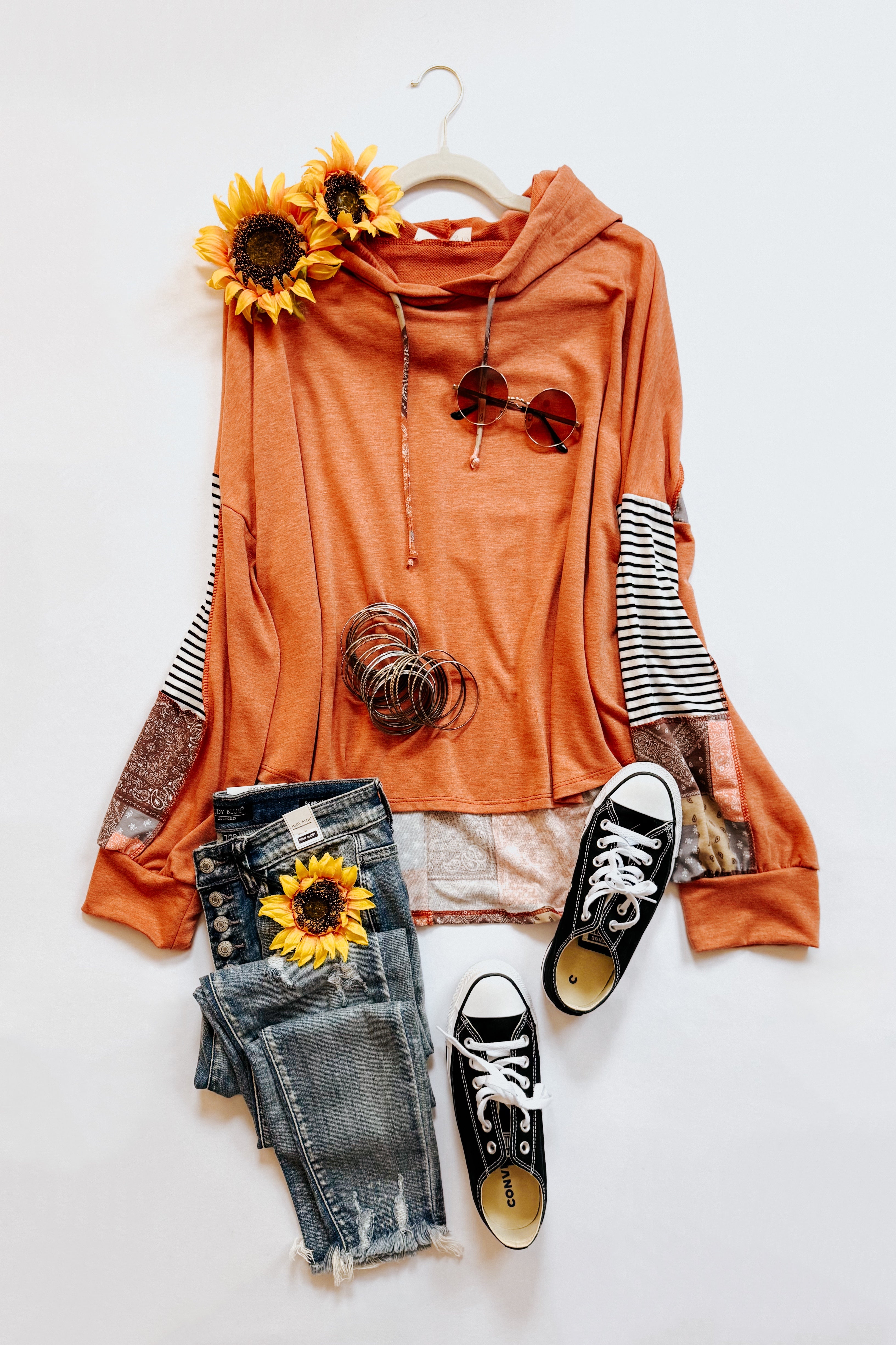 Rust hoodie with striped and patchwork sleeves paired with distressed denim jeans and black sneakers styled with silver bangles and sunflowers