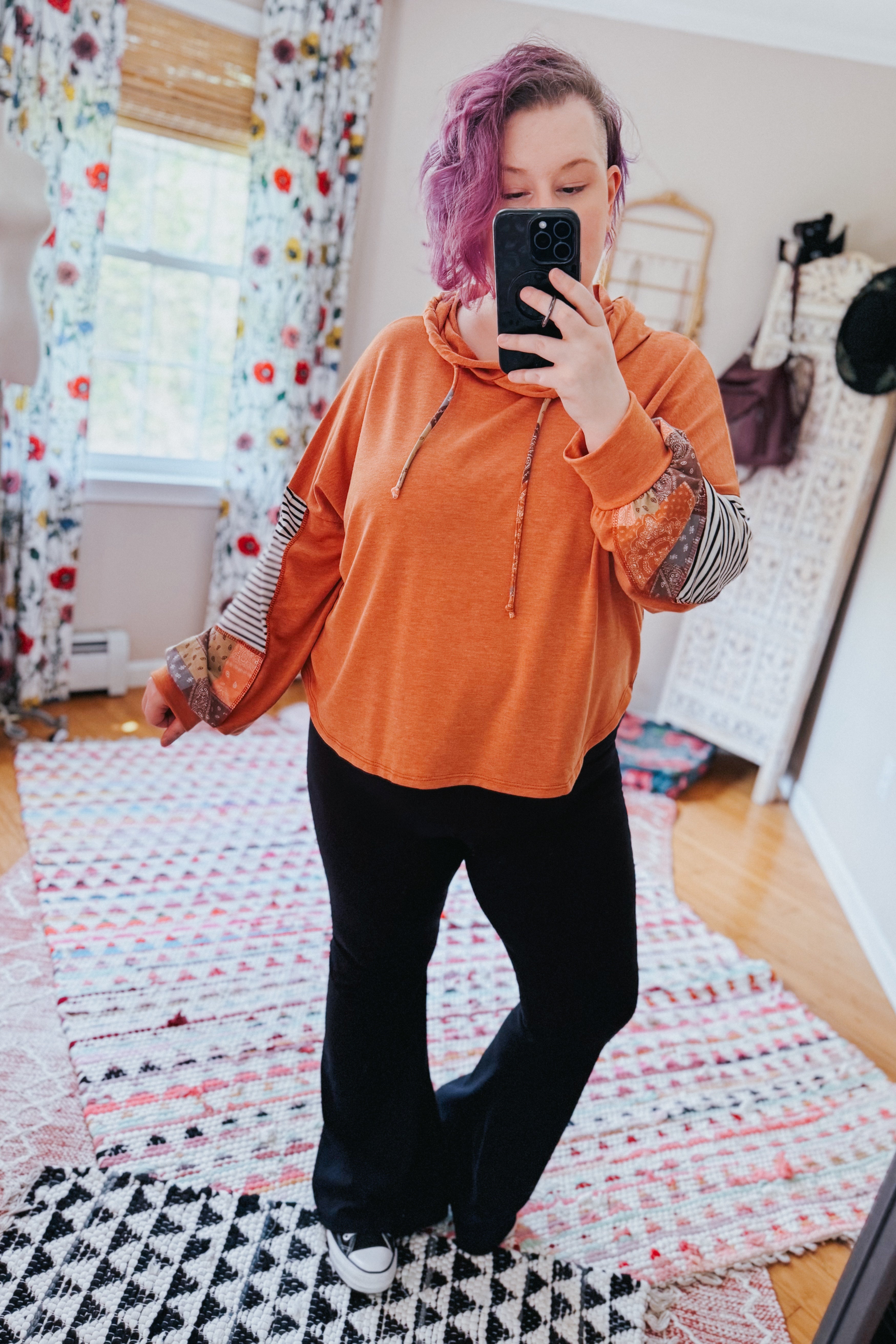 Woman with purple hair wearing rust hoodie with eclectic patchwork sleeves and black pants taking indoor selfie