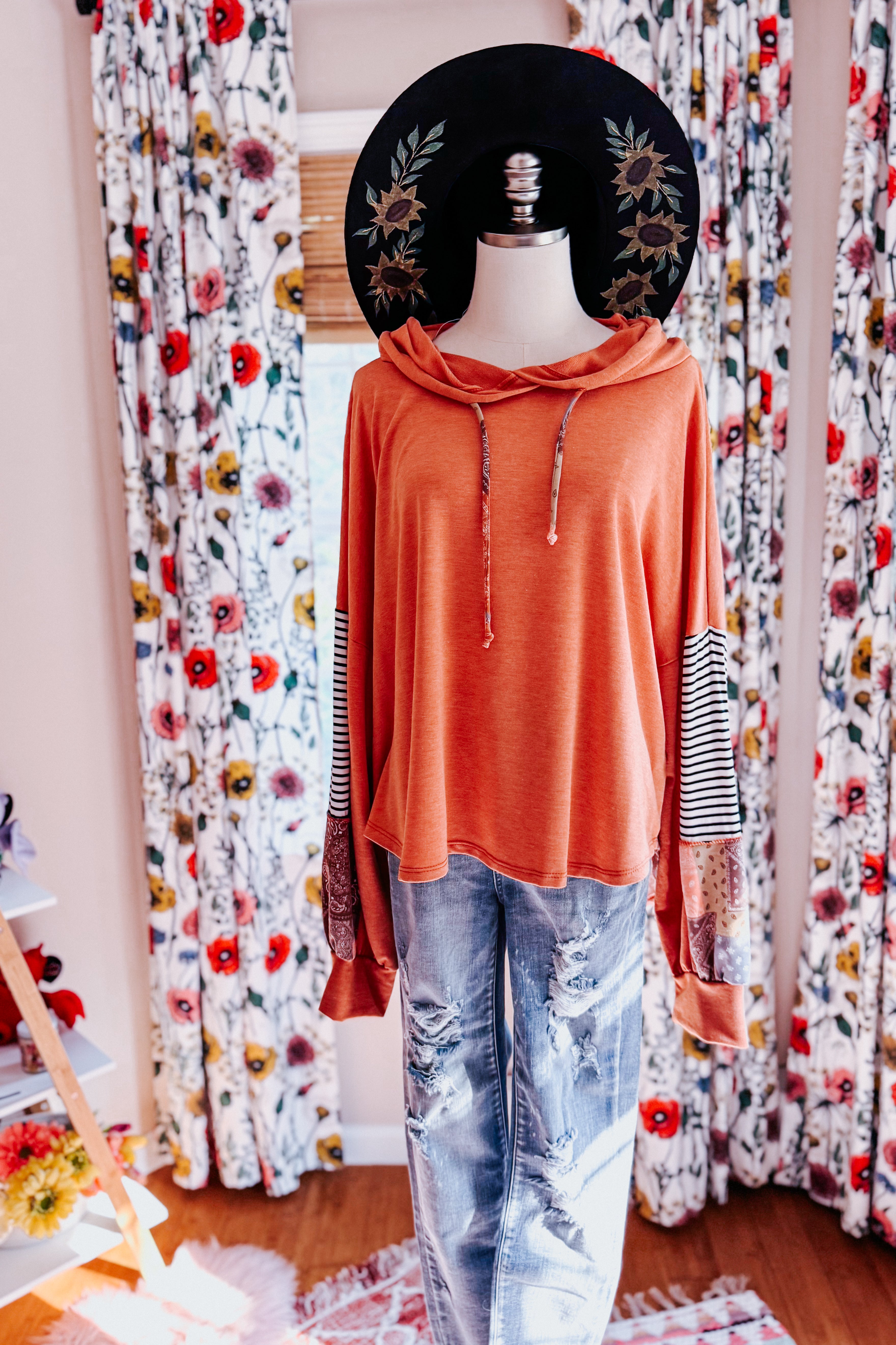 Rust-colored hoodie with mixed patterned sleeves paired with distressed light-wash jeans and floral black hat on mannequin