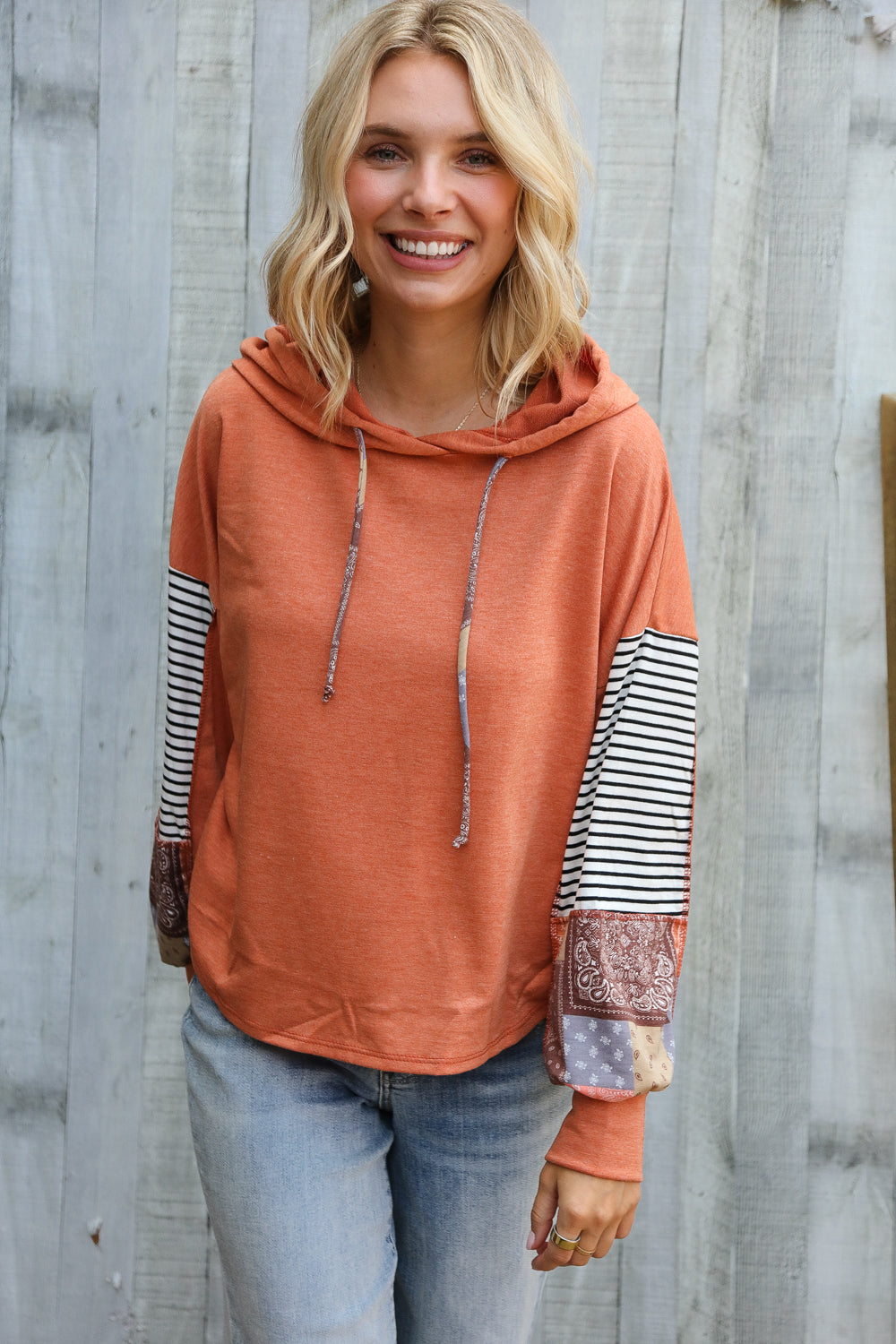 Smiling woman wearing rust hoodie with striped and patterned patchwork sleeves posing against weathered wooden wall