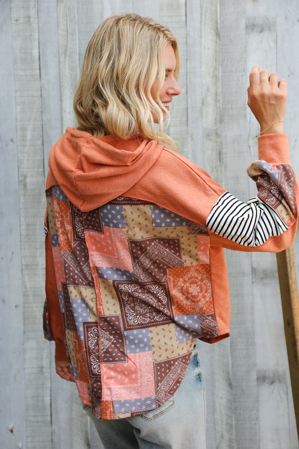Woman wearing rust hoodie with patchwork and striped sleeves posing sideways against weathered wooden wall