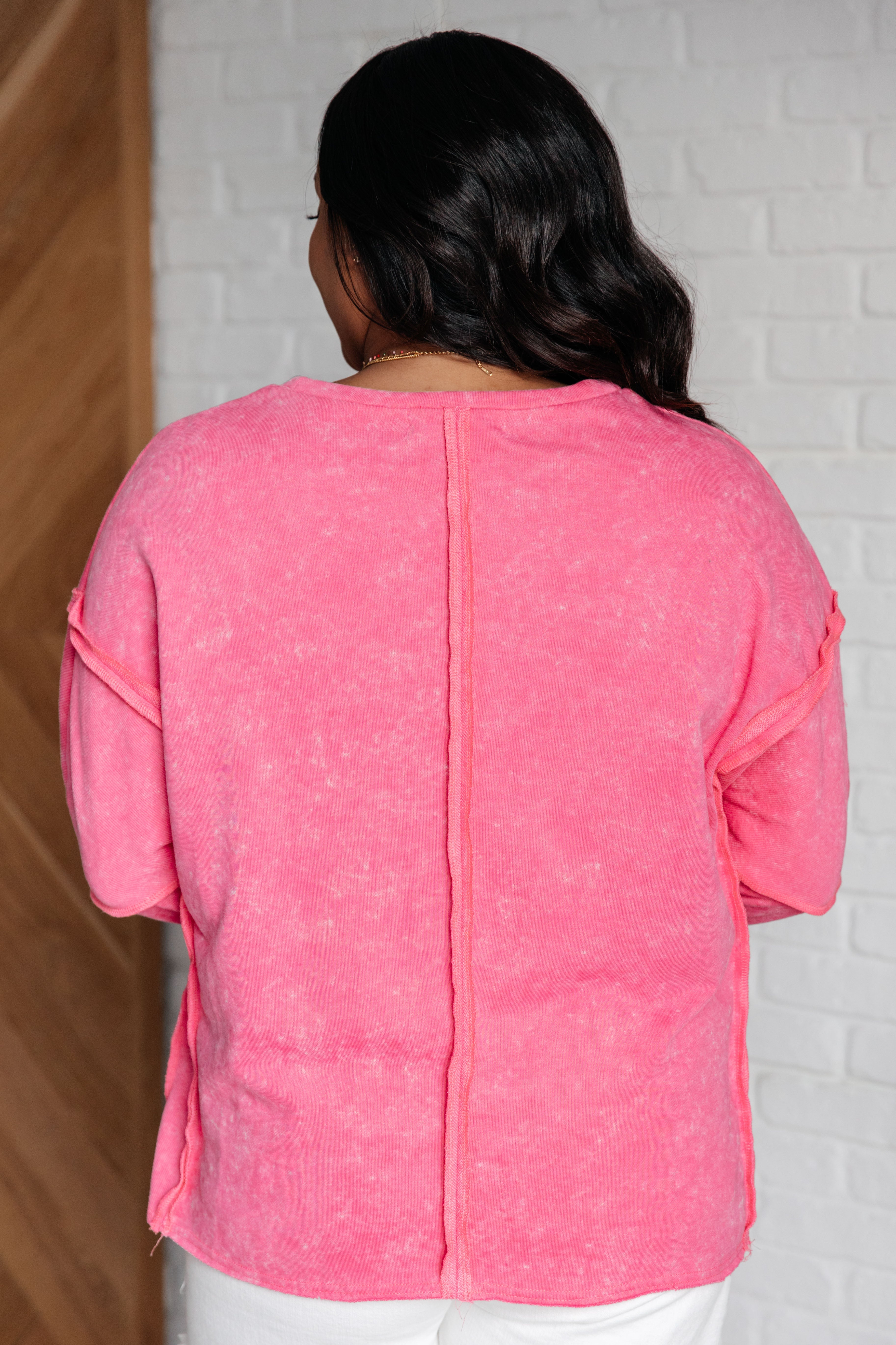 Woman wearing a pink mineral wash long-sleeve sweatshirt with exposed seams and white jean shorts standing against a white brick wall.