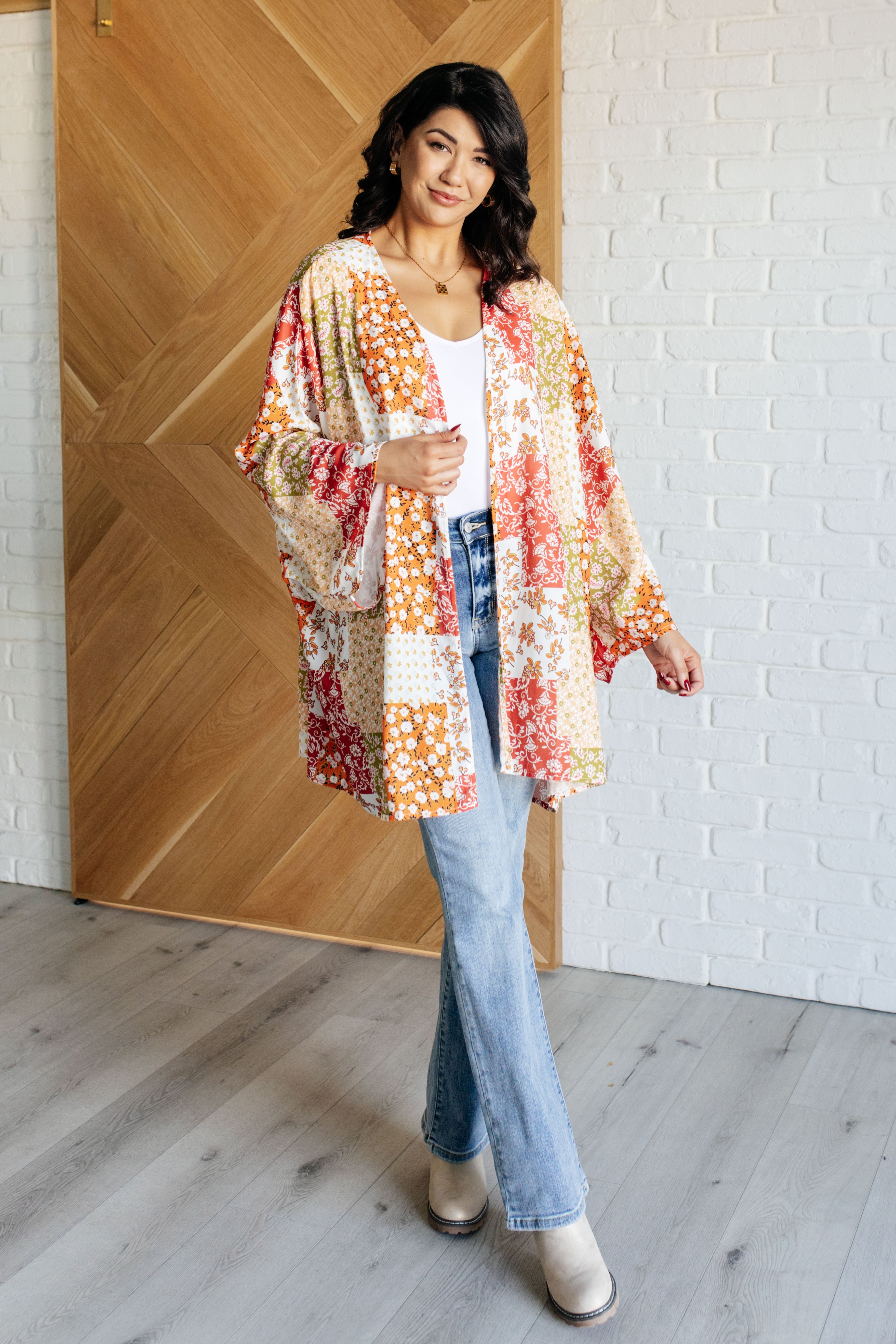 Woman wearing colorful orange and yellow mixed floral patchwork duster over white tank top and light blue jeans indoors by wooden door