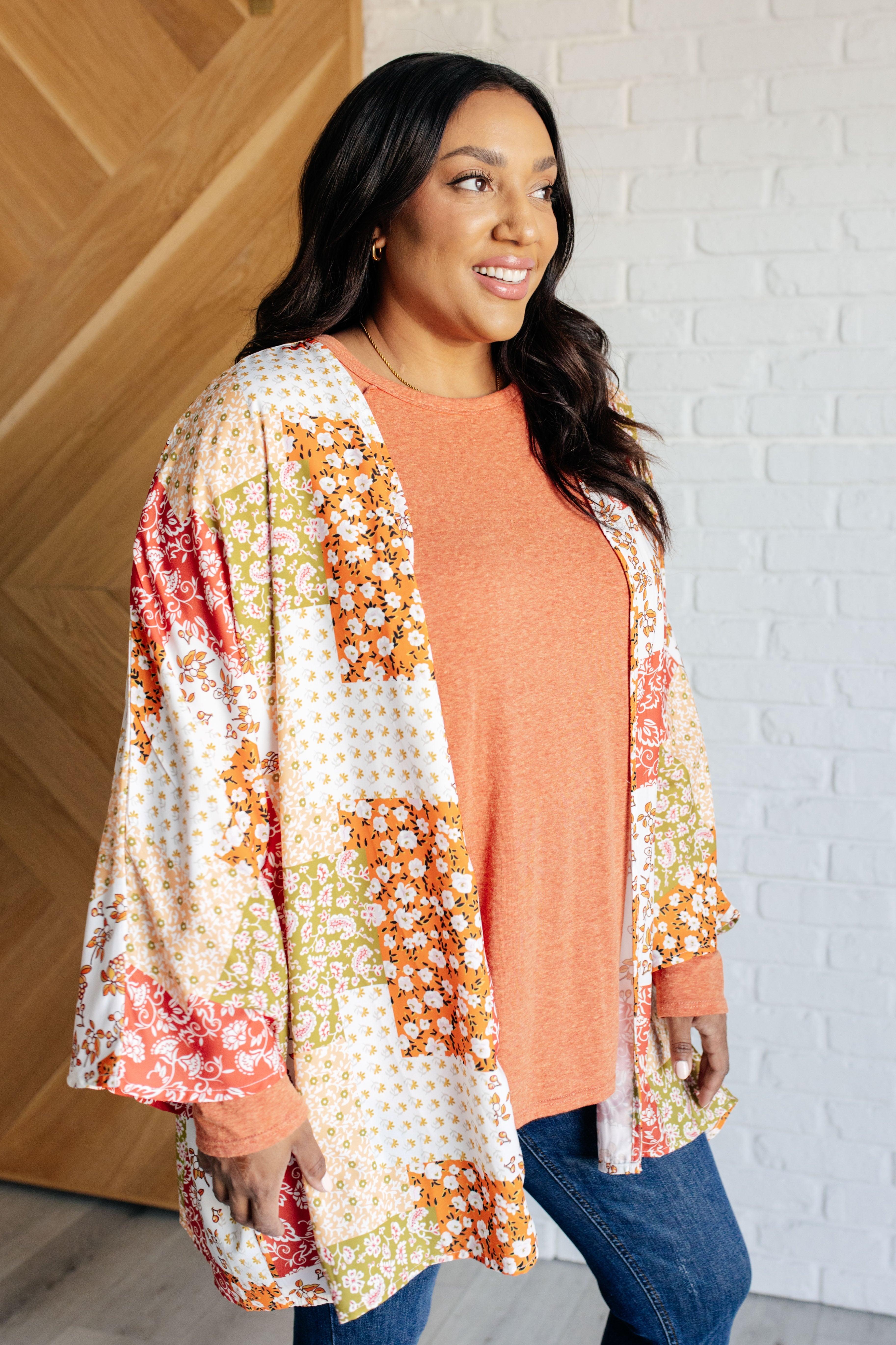 Woman smiling wearing orange and yellow mixed floral patchwork duster over orange long sleeve top indoors by white brick wall