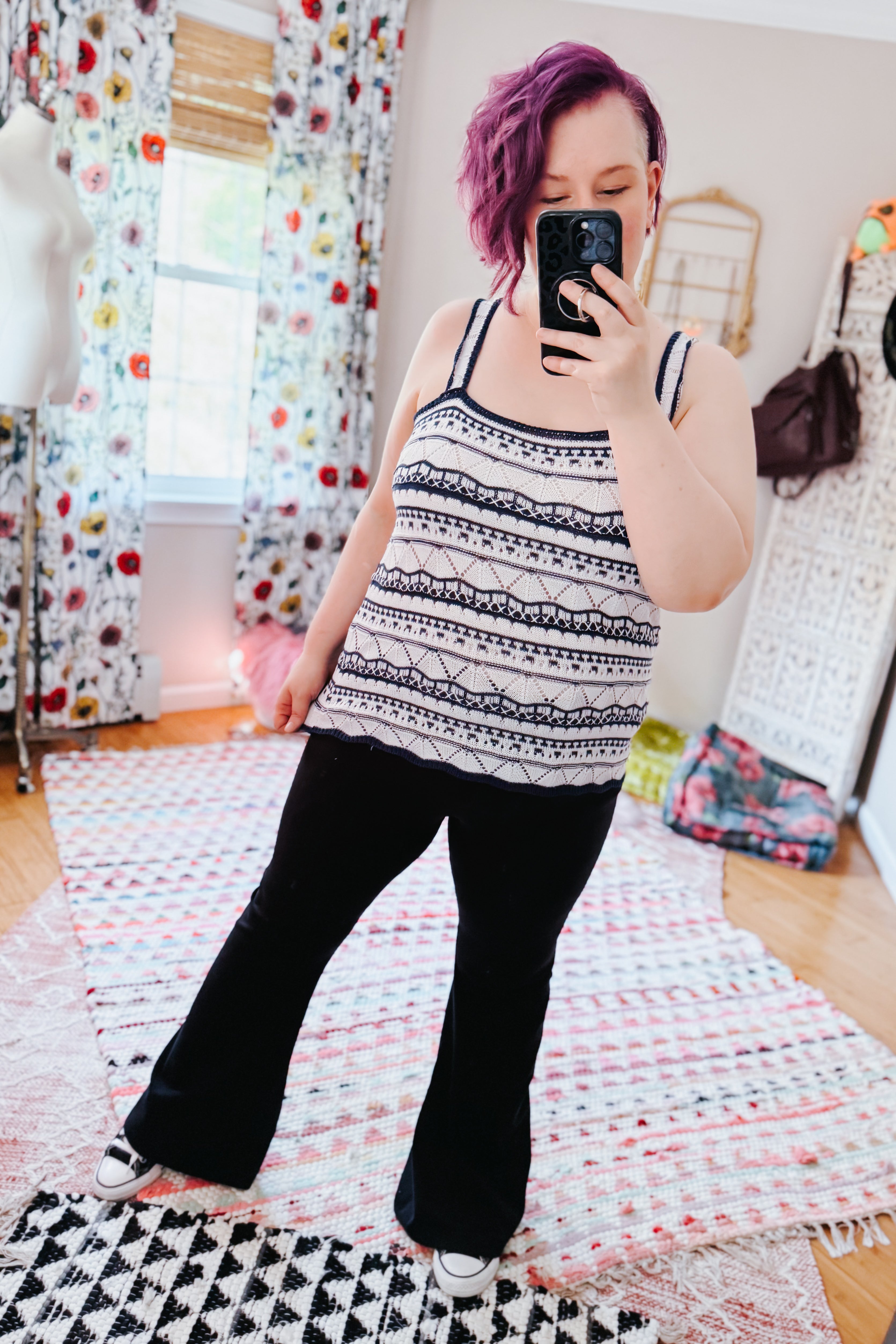 Woman with purple hair wearing a patterned knit tank with double straps, paired with black flared pants in a cozy, eclectic room