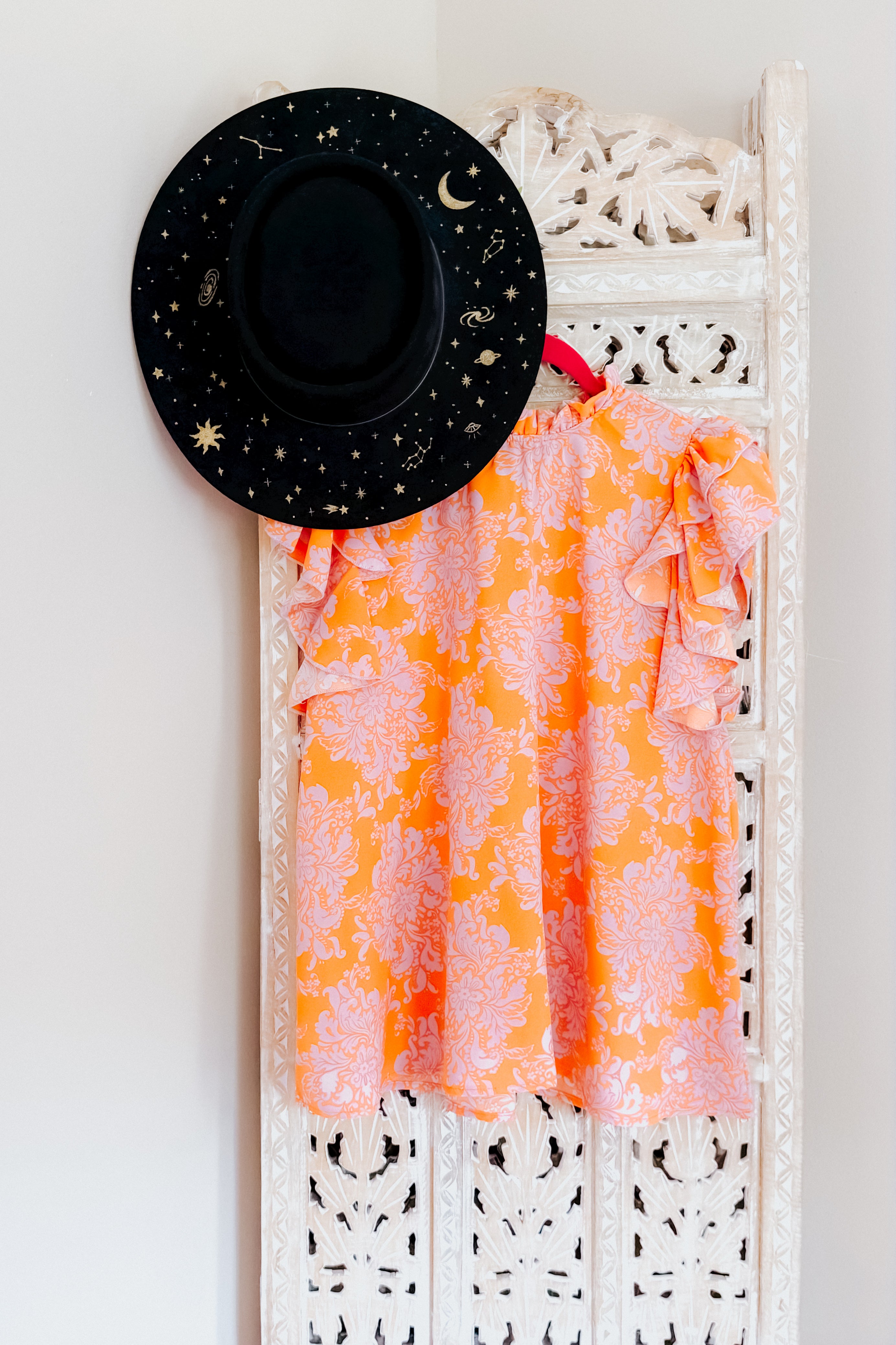Orange paisley patterned flutter sleeve top hanging next to black hat with gold celestial designs on carved white panel
