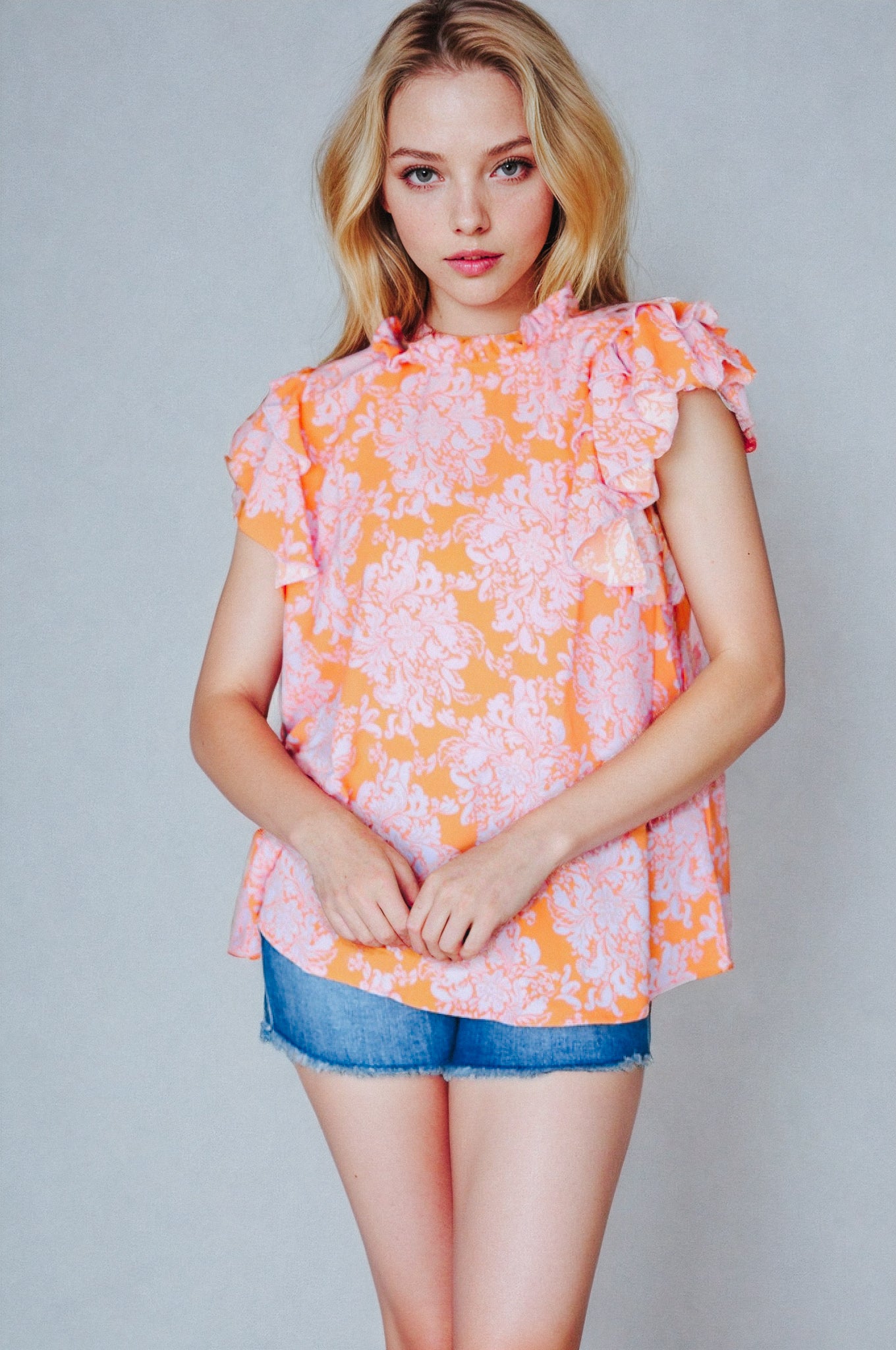 Woman wearing orange paisley mock neck top with flutter sleeves paired with blue denim shorts against gray background