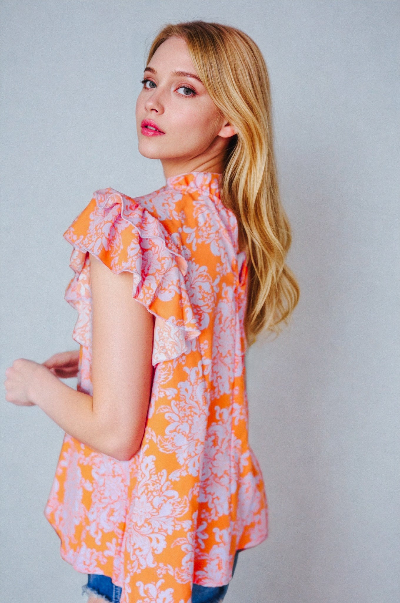Woman with long blonde hair wearing an orange paisley flutter sleeve top with mock neck against plain background