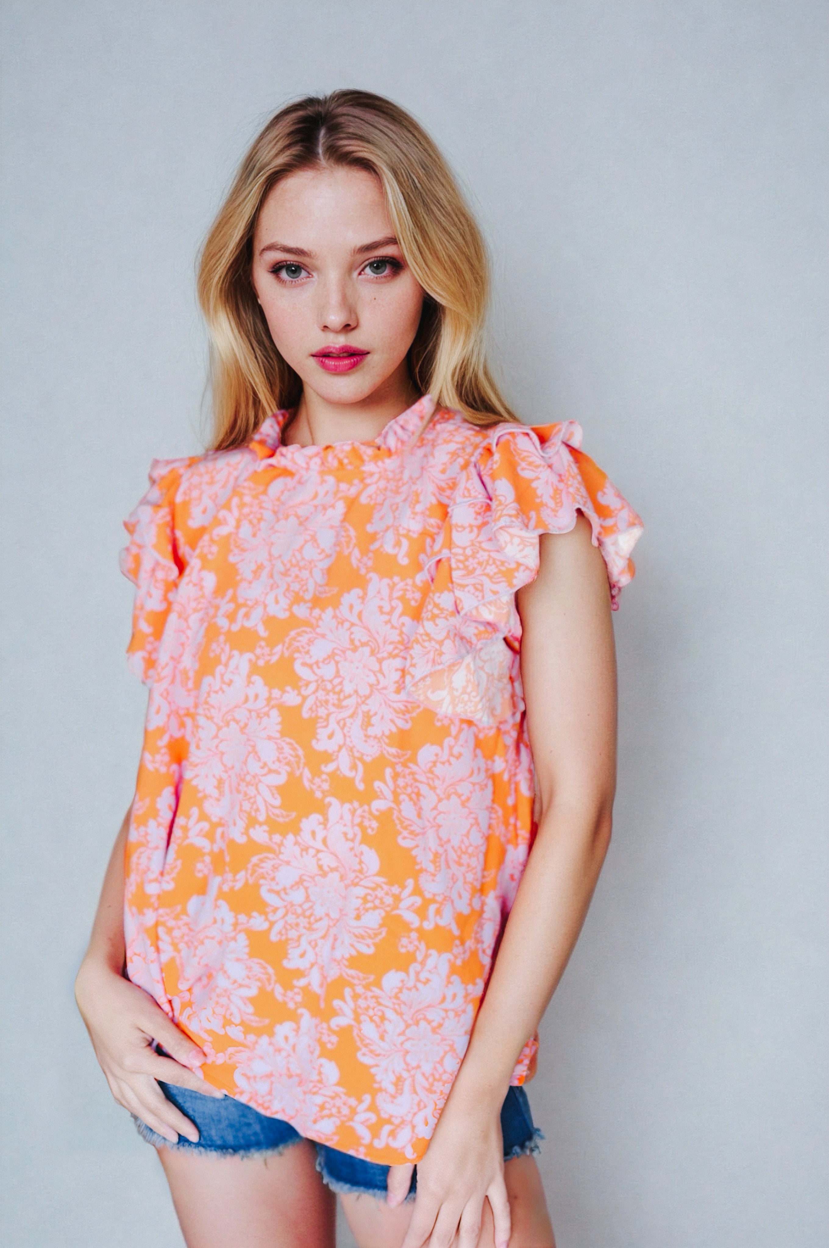 Young woman wearing orange paisley mock neck top with flutter sleeves and denim shorts against gray background