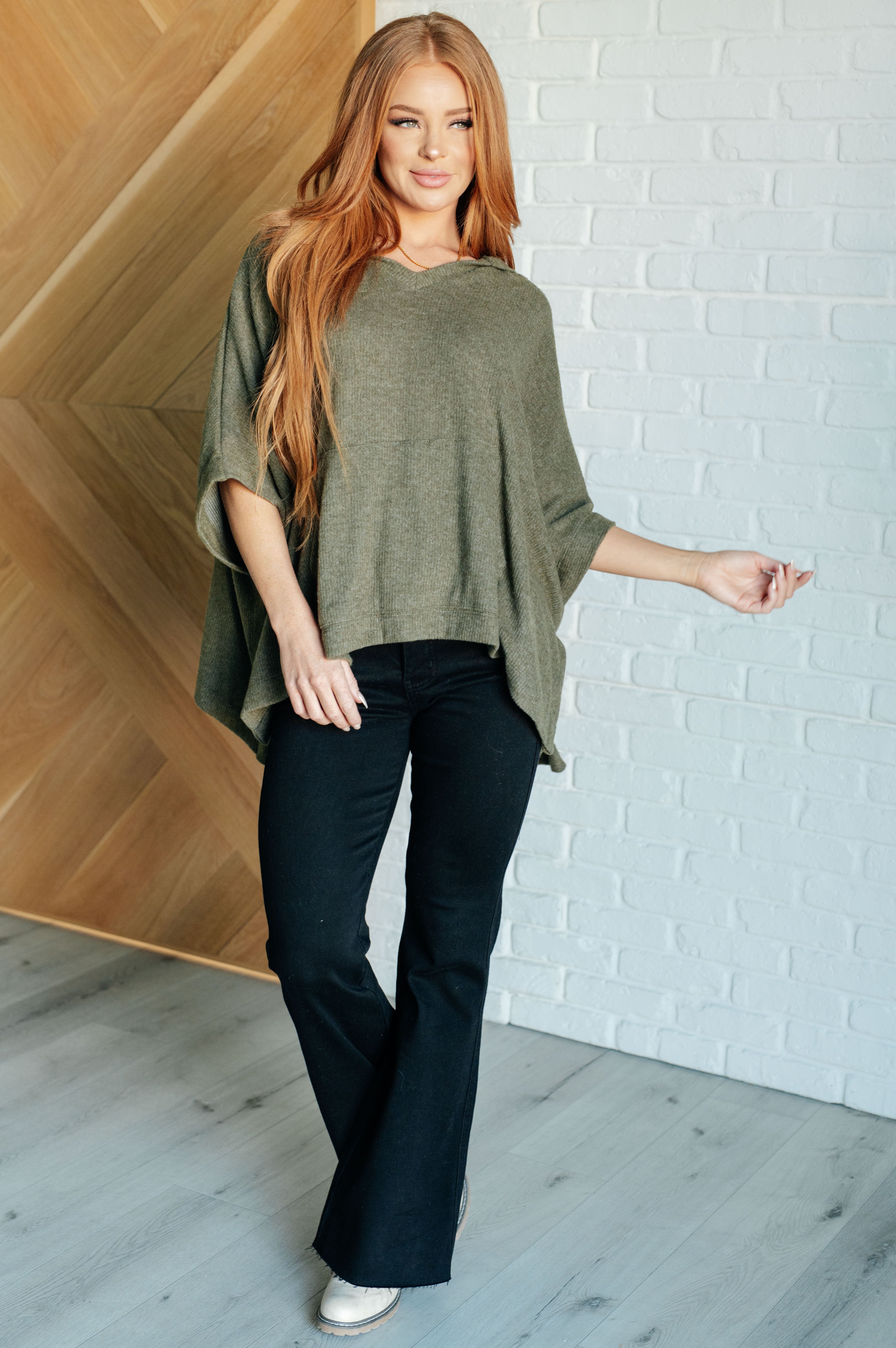 Woman wearing an olive hooded poncho with loose fit styled with black flare pants and white boots indoors