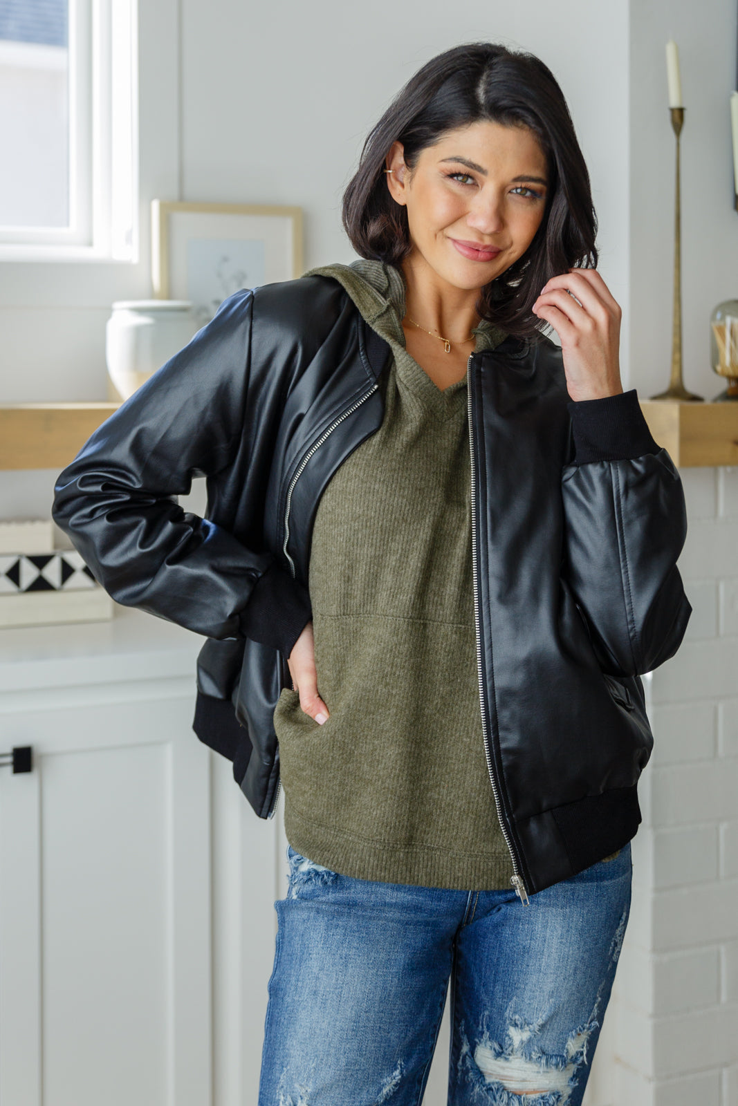 Woman wearing an olive knit hooded top layered under a black leather jacket, styled with ripped blue jeans indoors