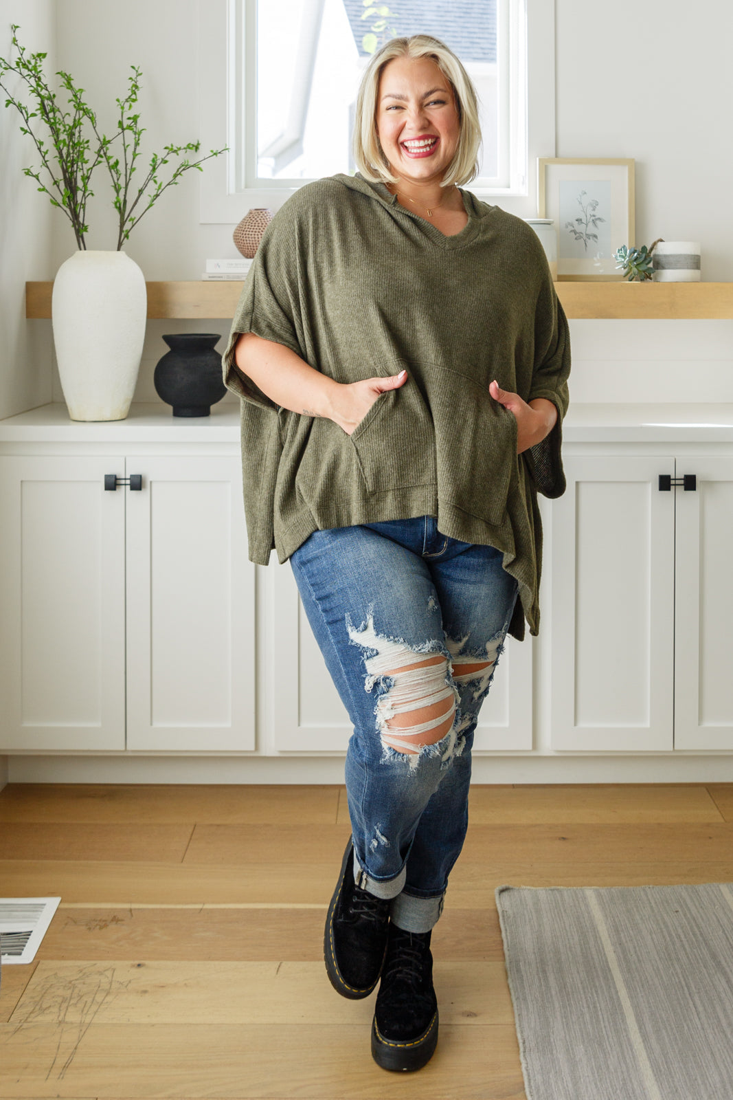 Smiling plus-size woman wearing an olive hooded poncho with pockets paired with ripped jeans and black shoes indoors