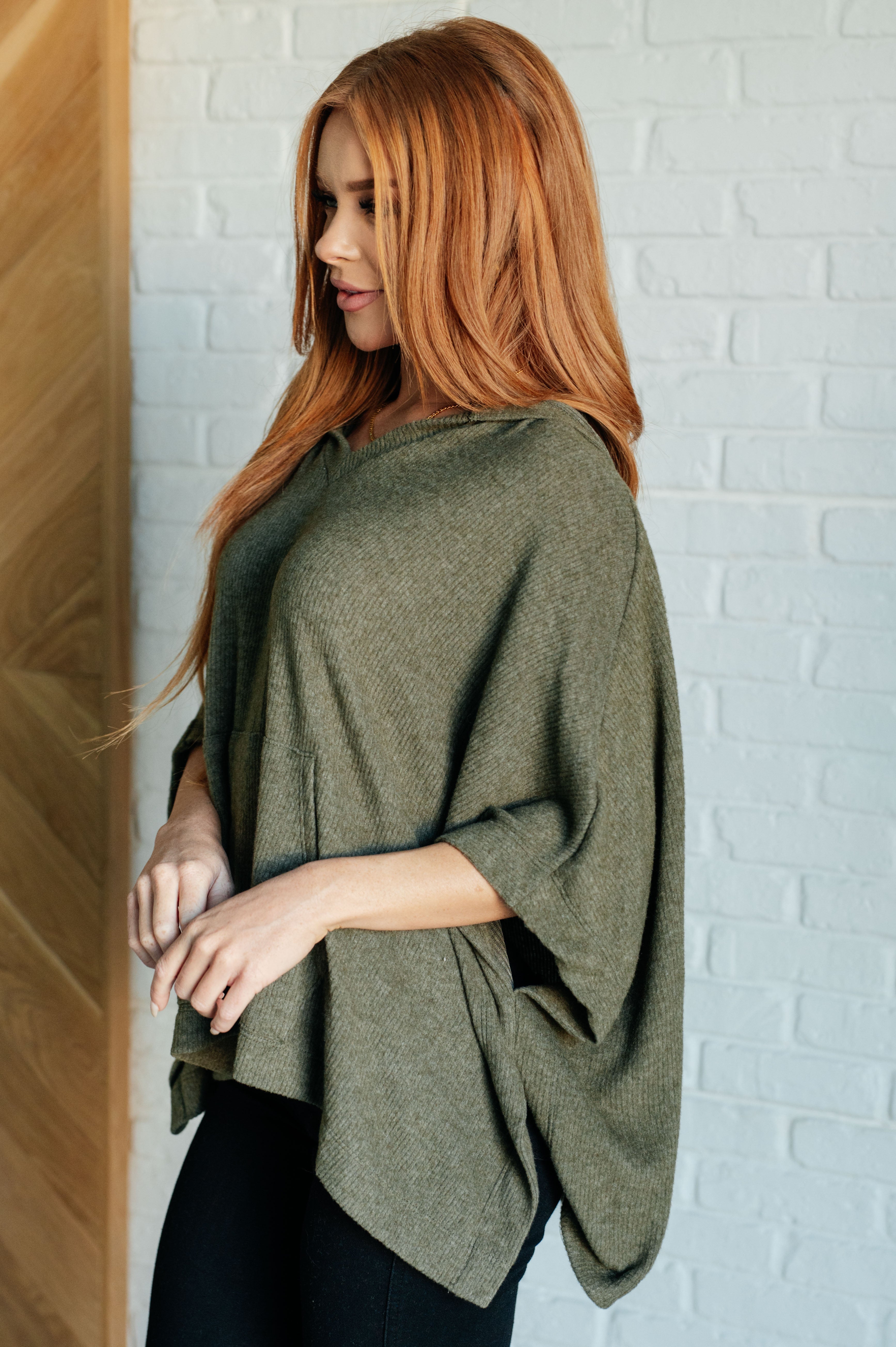 Woman with red hair wearing an olive knit poncho with hood and front pocket, styled with black pants indoors