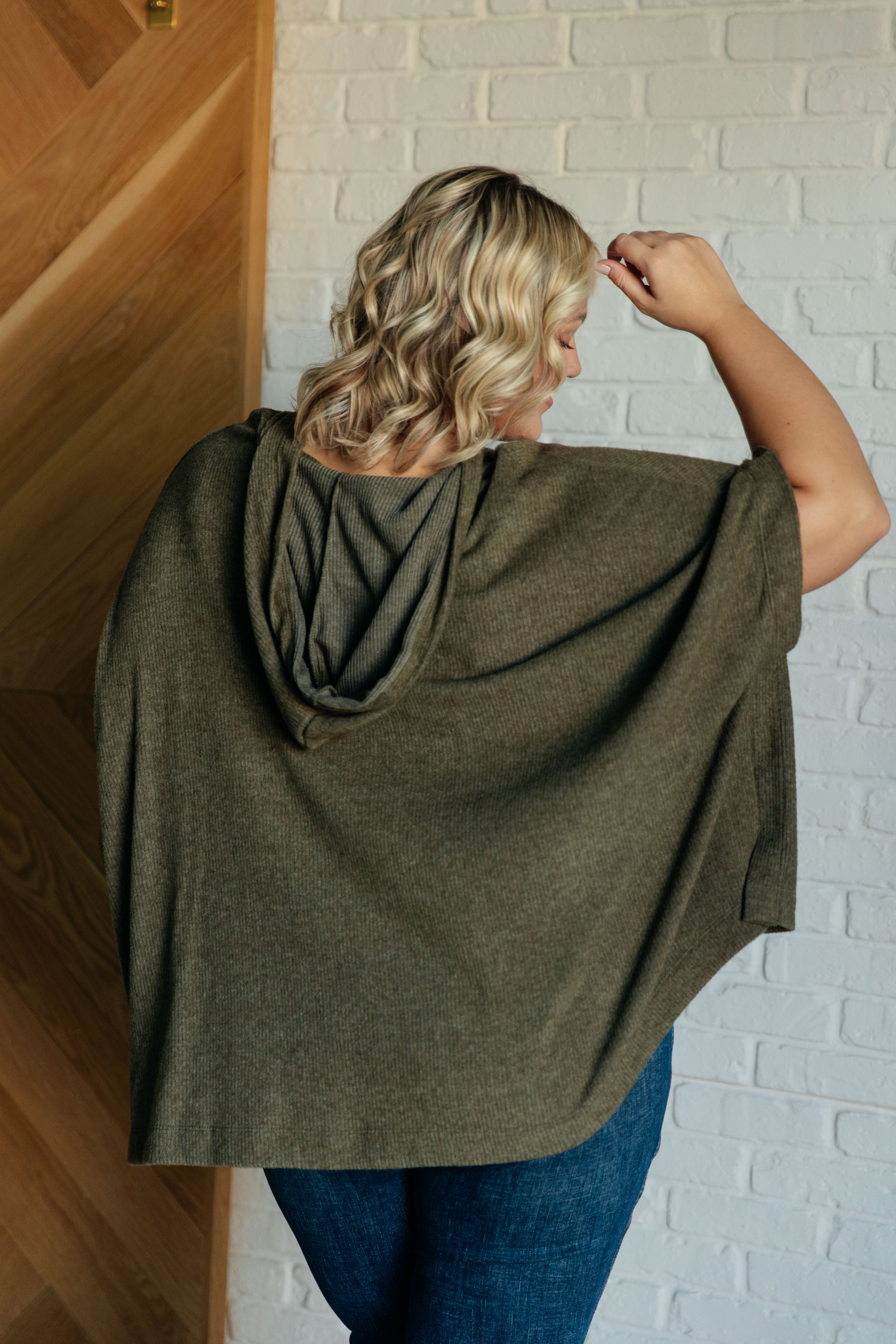 Plus-size woman with wavy blonde hair wearing an olive hooded poncho with loose fit, shown from the back indoors