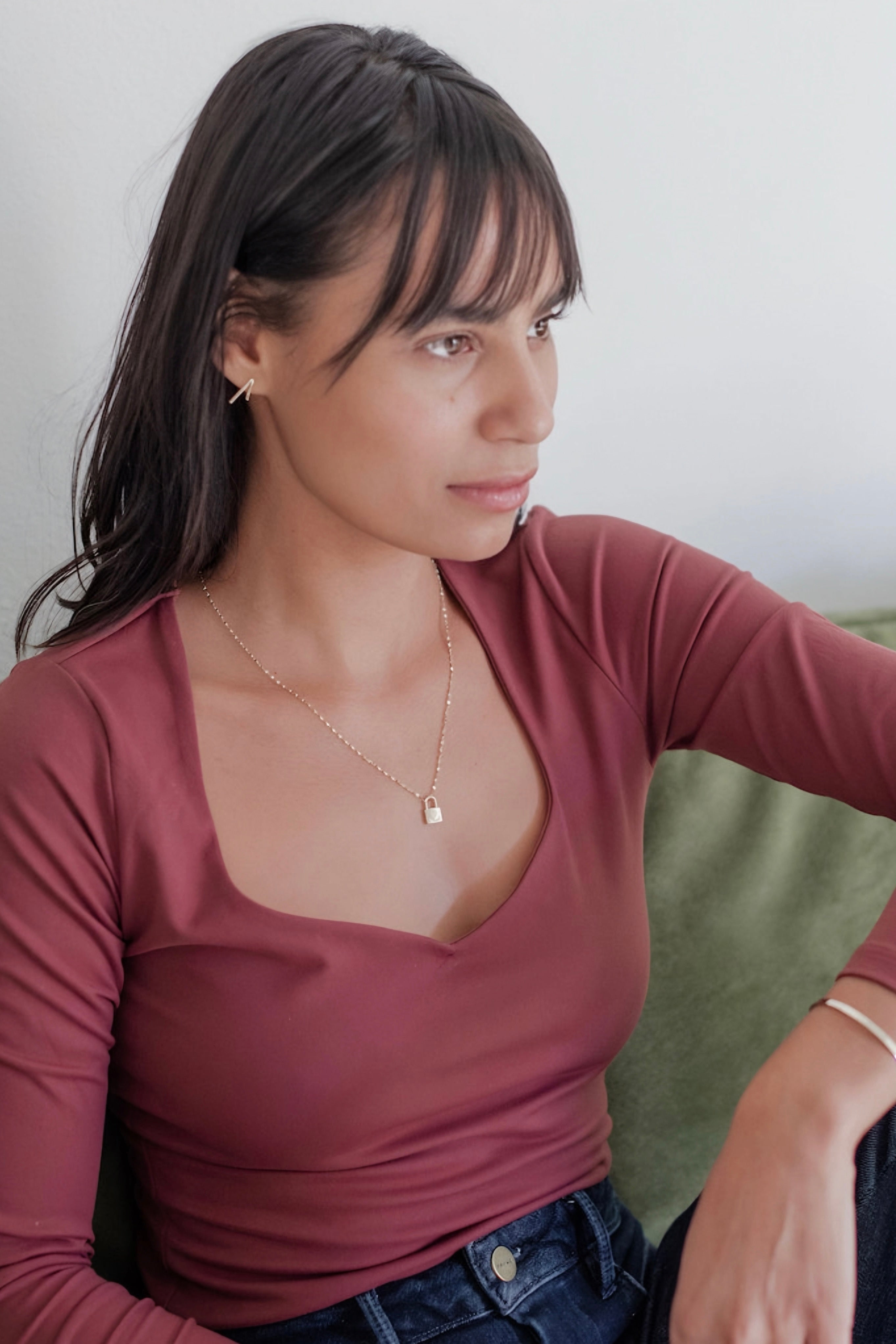 Woman wearing gold love locked necklace with delicate chain and mauve scoop neck top seated indoors