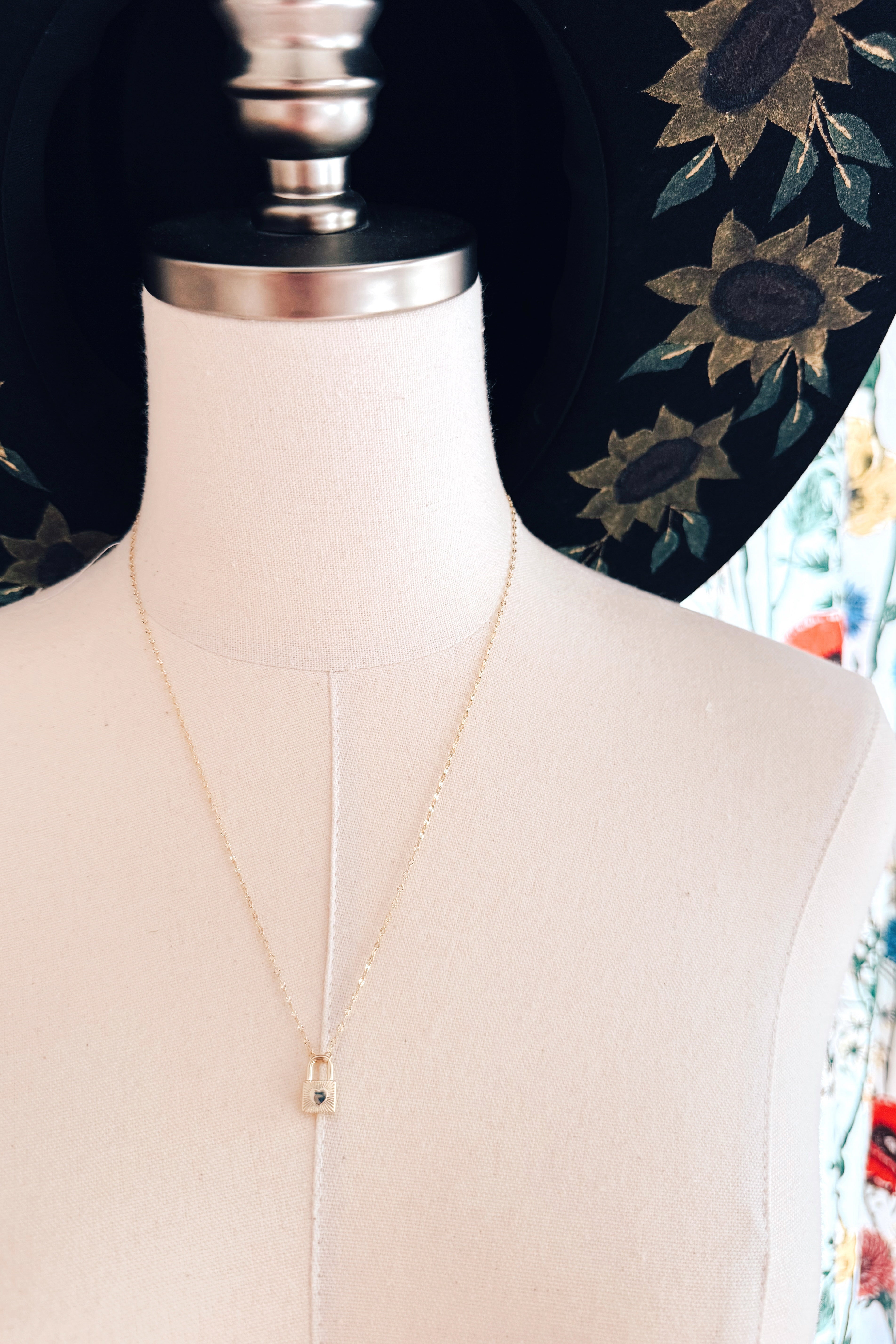 Delicate gold chain with small lock pendant displayed on fabric mannequin wearing sunflower embroidered black hat in eclectic style