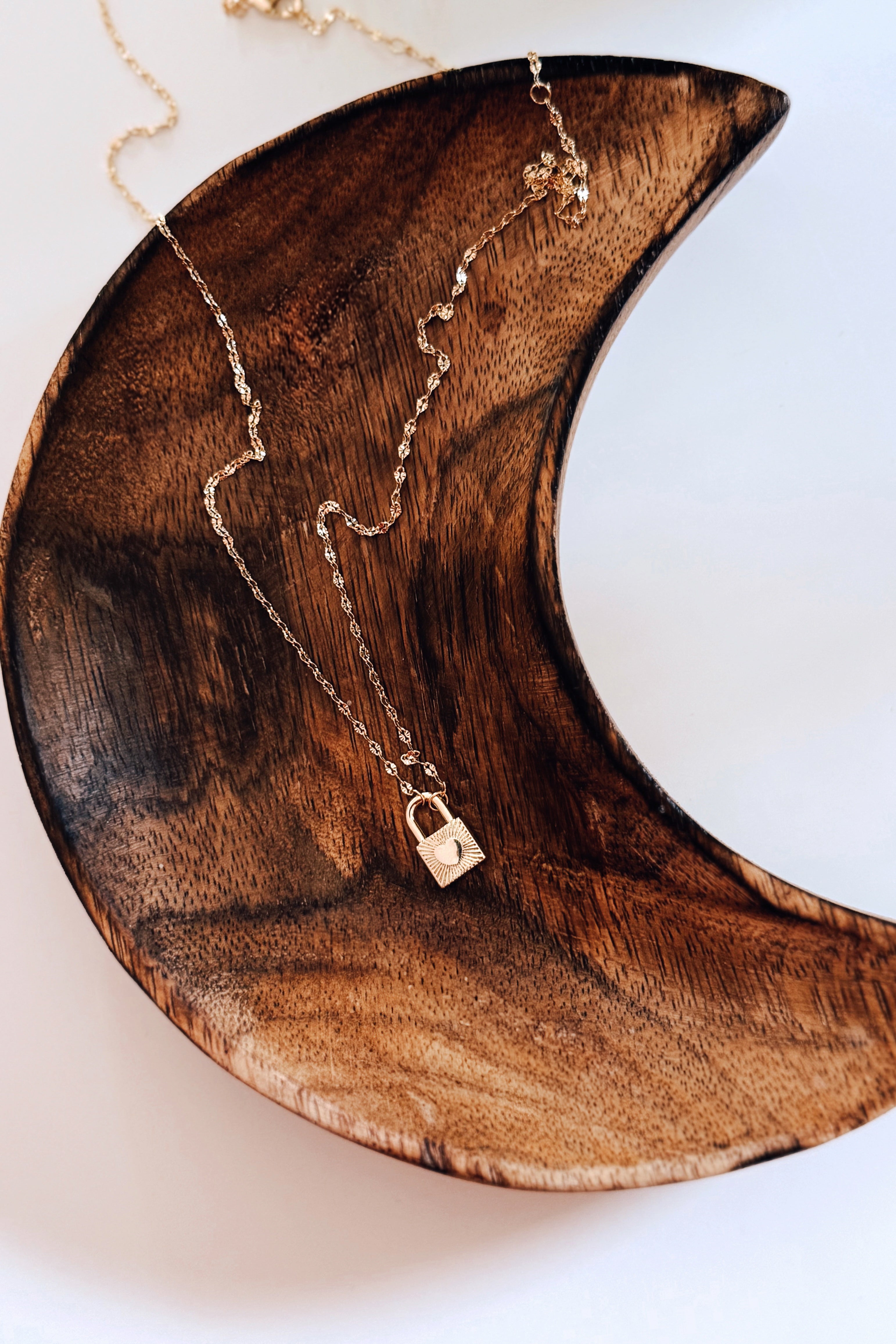 Gold delicate chain with small padlock pendant resting on curved dark wood dish in bold, free-spirited style