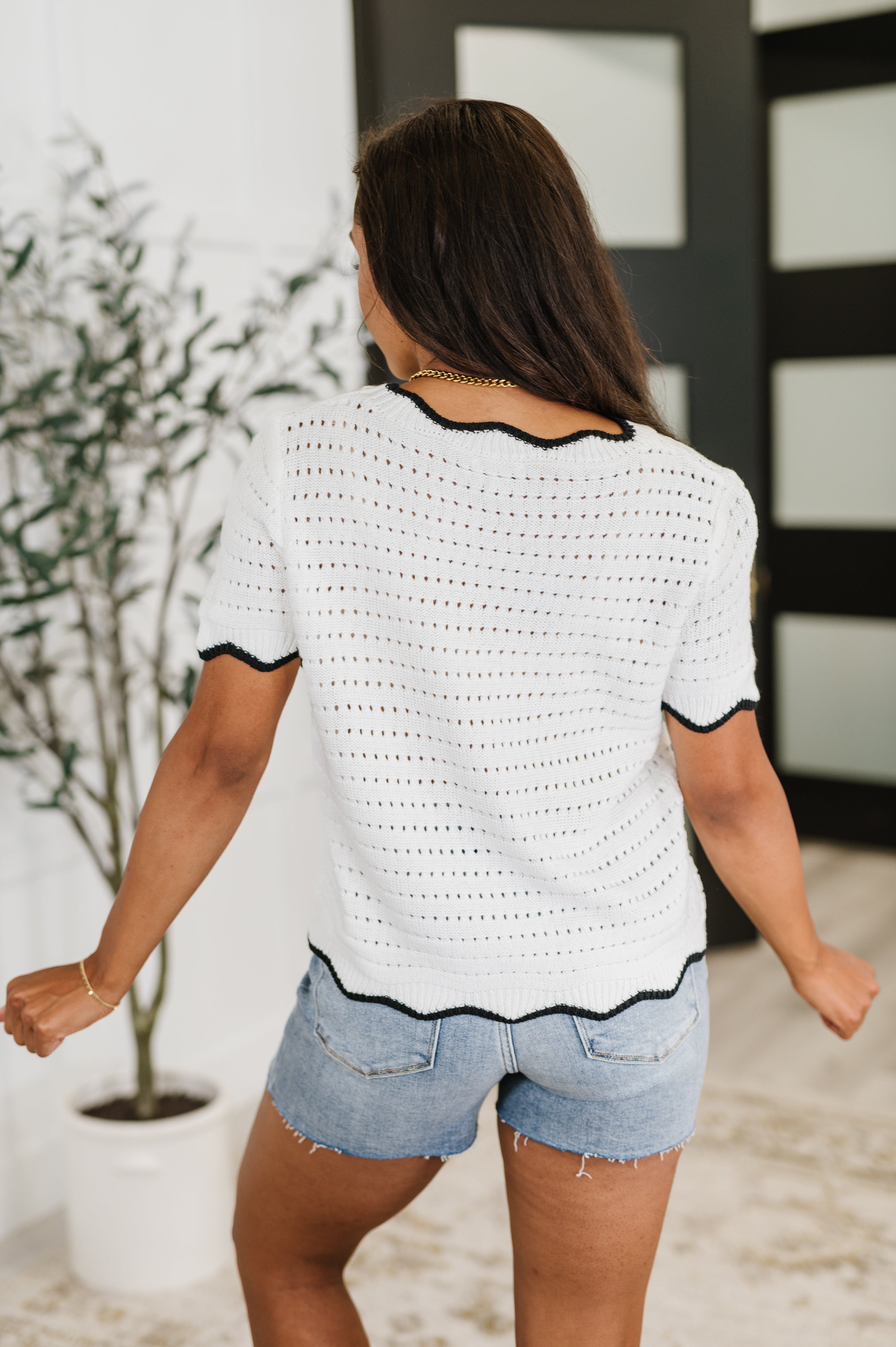 Back view of woman wearing a white wavy eyelet knit top with black trim and casual denim shorts indoors, eclectic style