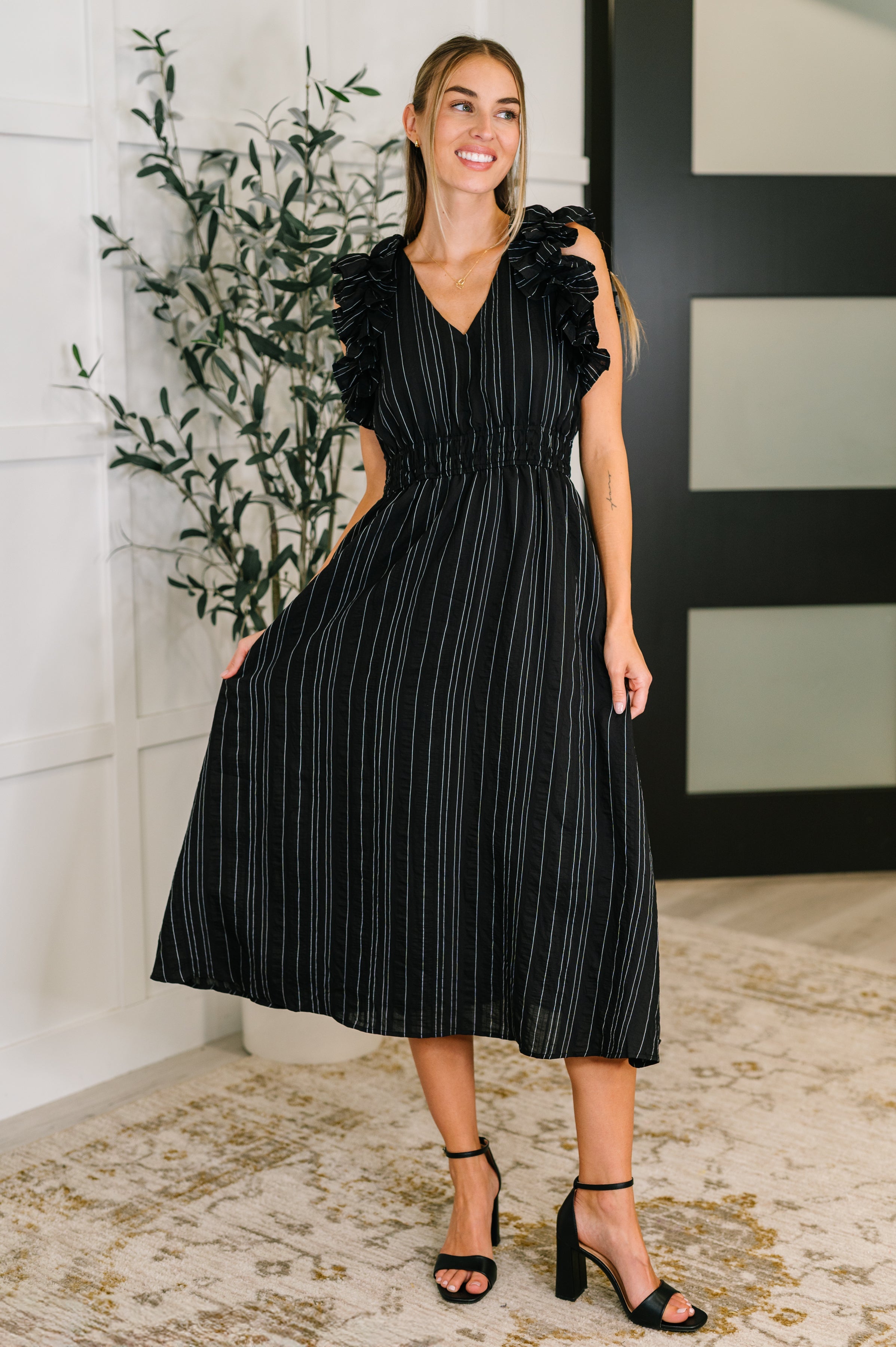 Woman wearing a black pinstripe dress with ruffled sleeves and V-neck standing indoors on patterned rug