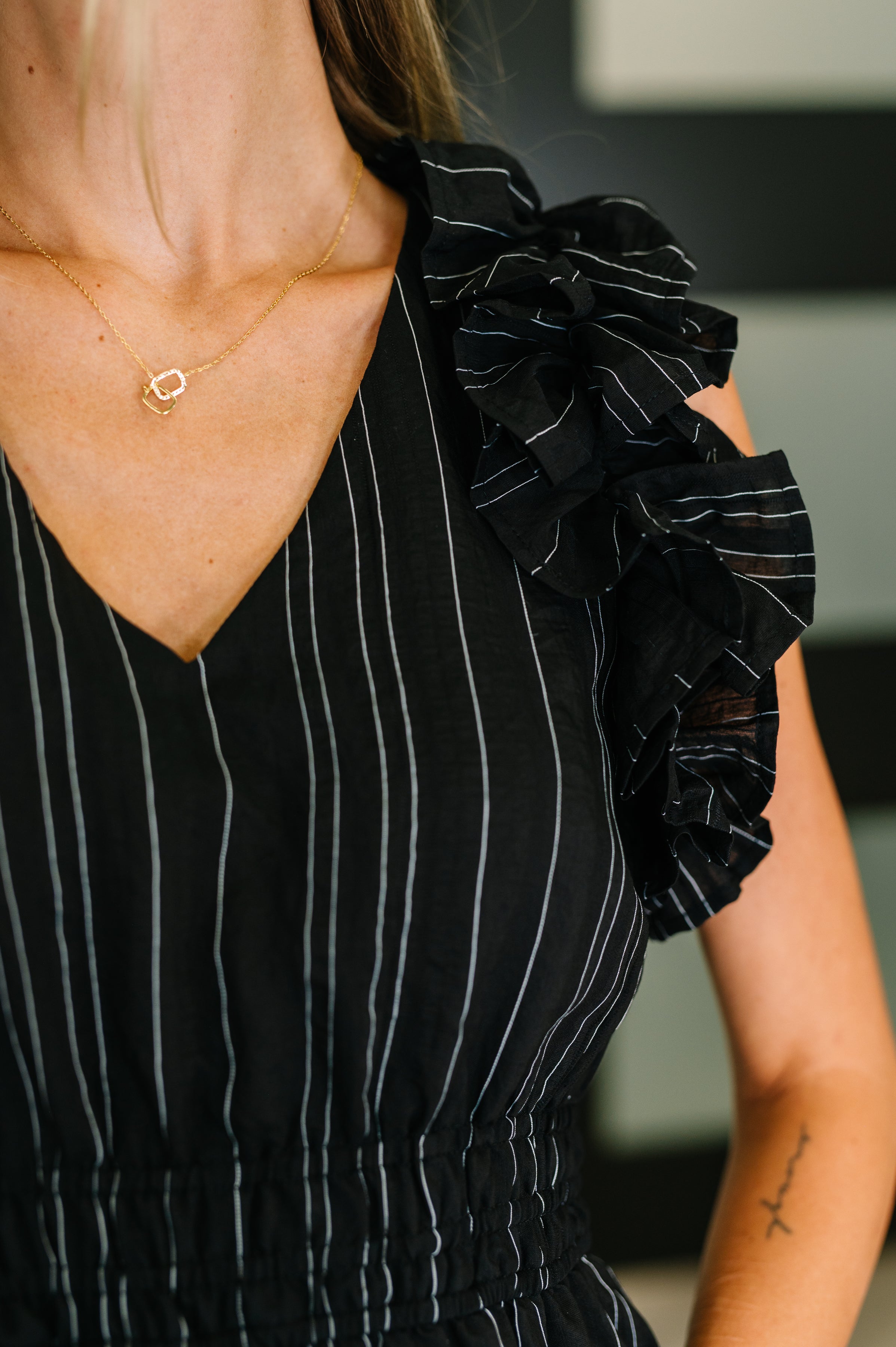 Close-up of a black pinstripe dress with V-neck and ruffled sleeve detail worn with a delicate gold necklace